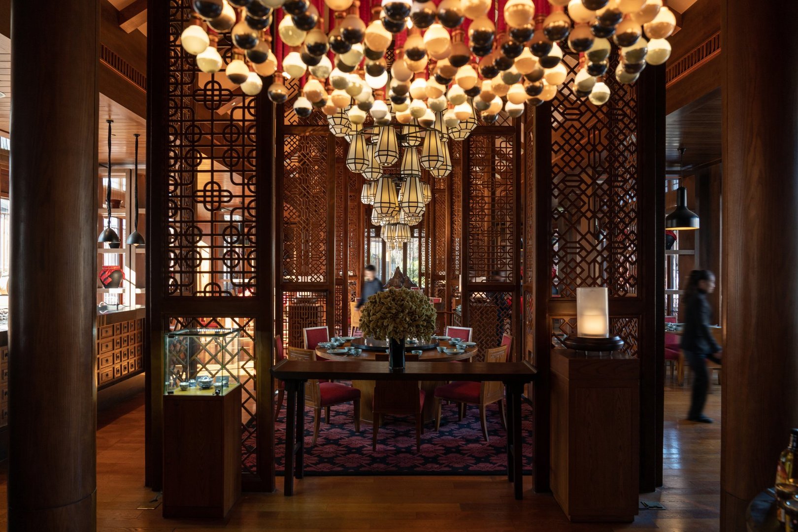 Elegant dining area in a traditional Chinese restaurant featuring intricate wooden lattice partitions, warm ambient lighting, round dining tables with red chairs, and ornate pendant lamps, ideal for showcasing Asian hospitality, interior design, and cultural dining experiences.