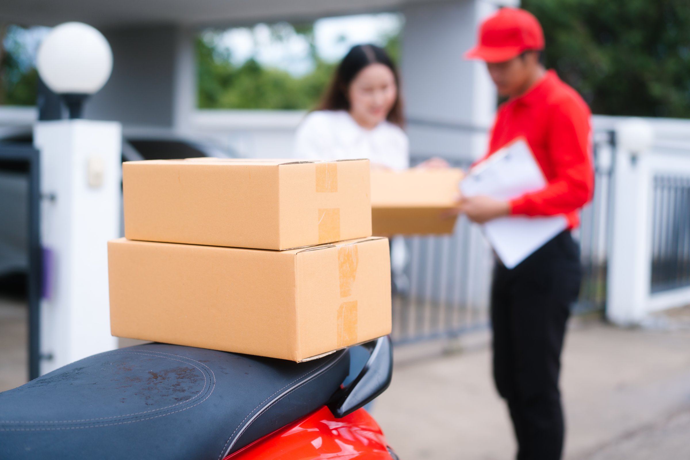 Attractive Asian delivery man in a red uniform hands a package to a middle-aged woman who is signing a clipboard next to a red motorcycle loaded with more boxes.