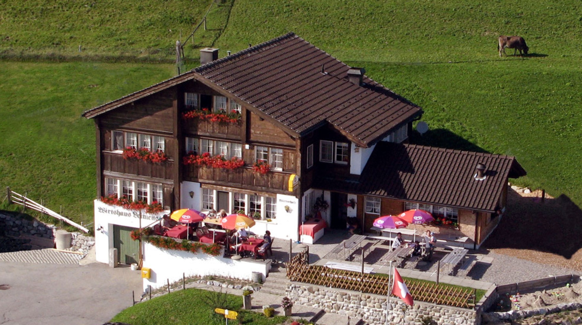 Rustic Swiss chalet with outdoor seating, flower boxes, and a cow on a grassy hill. Swiss flag is visible.