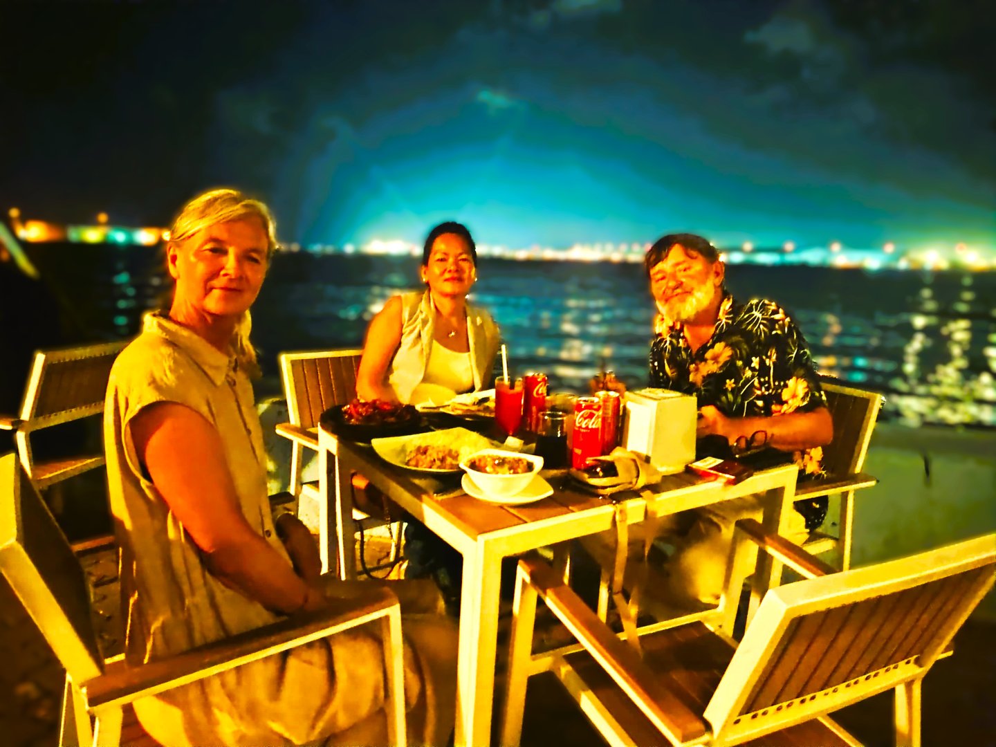 Three people sitting at a table by the beach at night, enjoying drinks and snacks with city lights in the background.