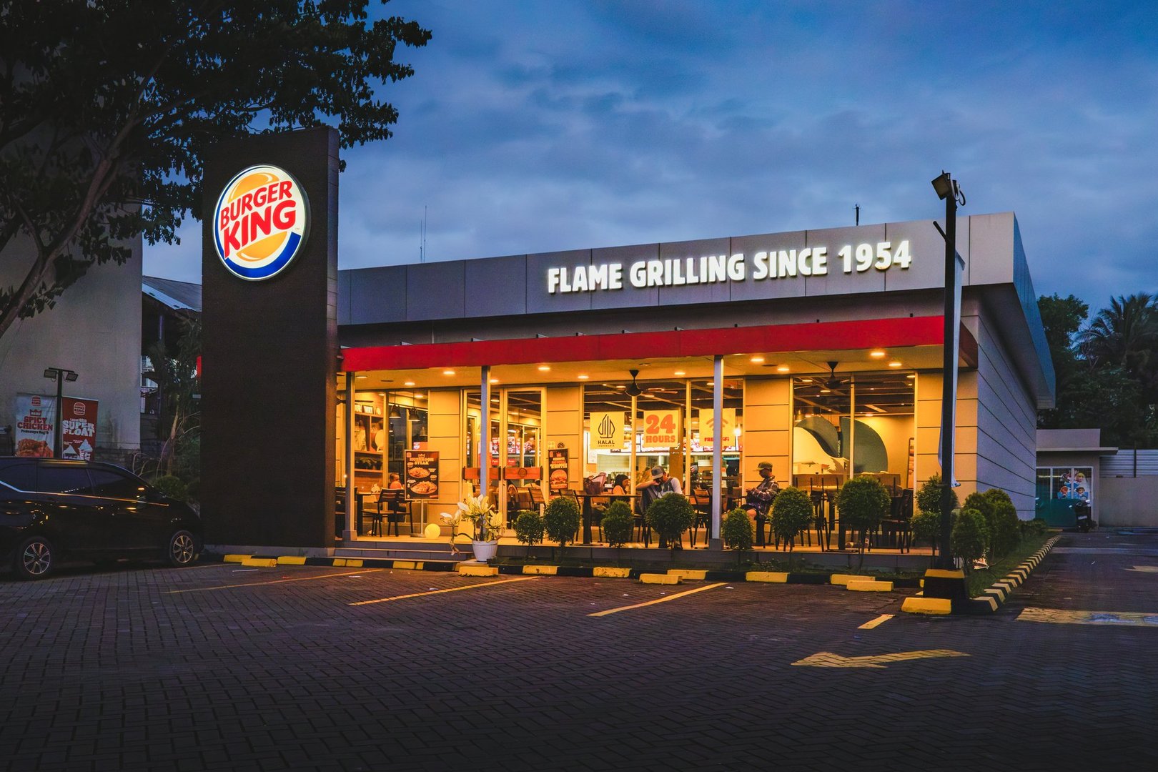 Balikpapan, Indonesia - Feb 22nd 2025. Exterior of a Burger King restaurant at dusk, with a dark vertical sign with the logo, and FLAME GRILLING SINCE 1954 text, and brightly lit interior visible through large windows. Trees and a paved parking lot are also visible.