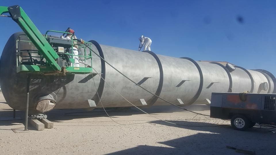 Workers in protective suits sandblast a large metal cylinder on a sunny day at an industrial site.