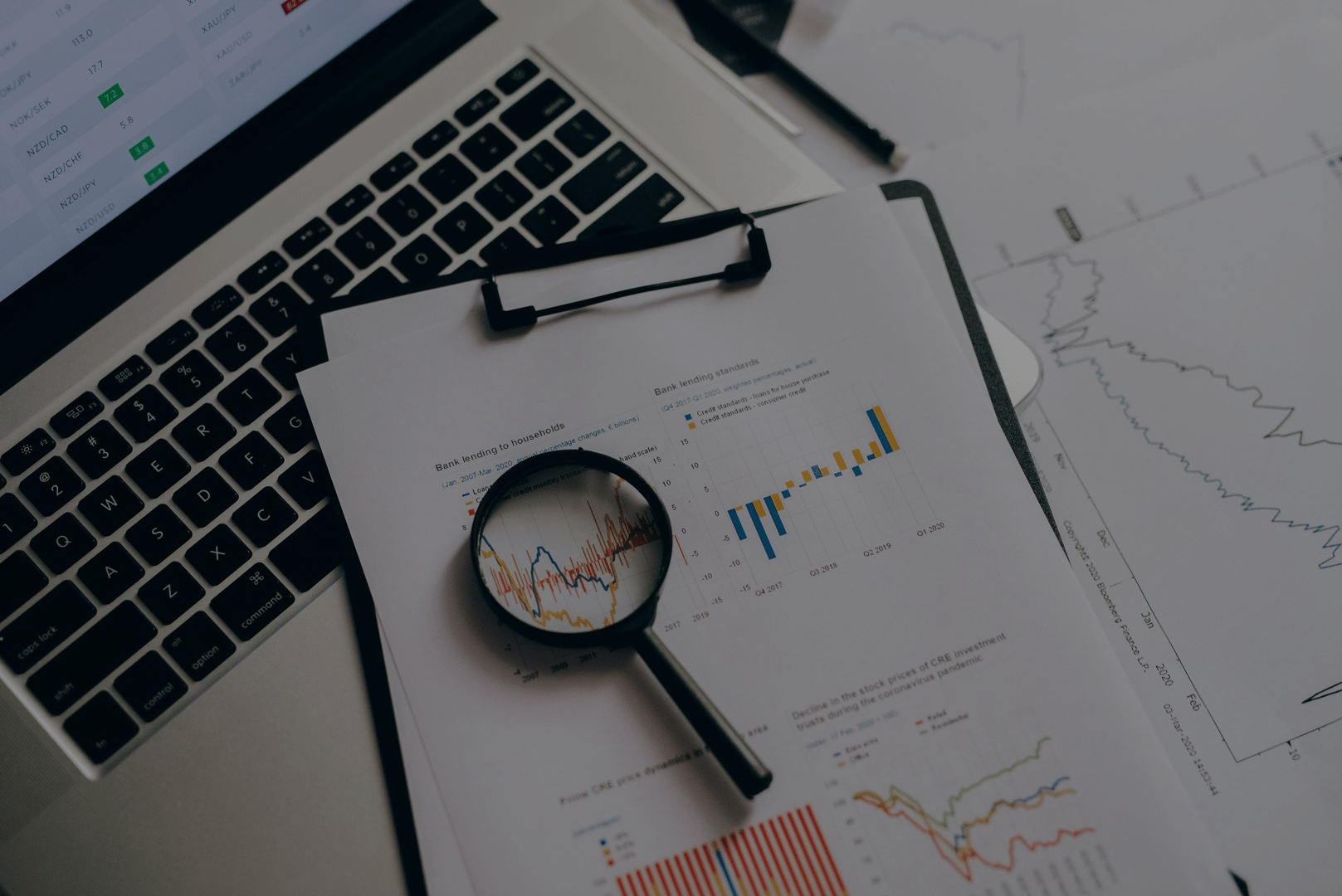 Magnifying glass on financial charts and graphs on a clipboard beside a laptop, indicating data analysis.