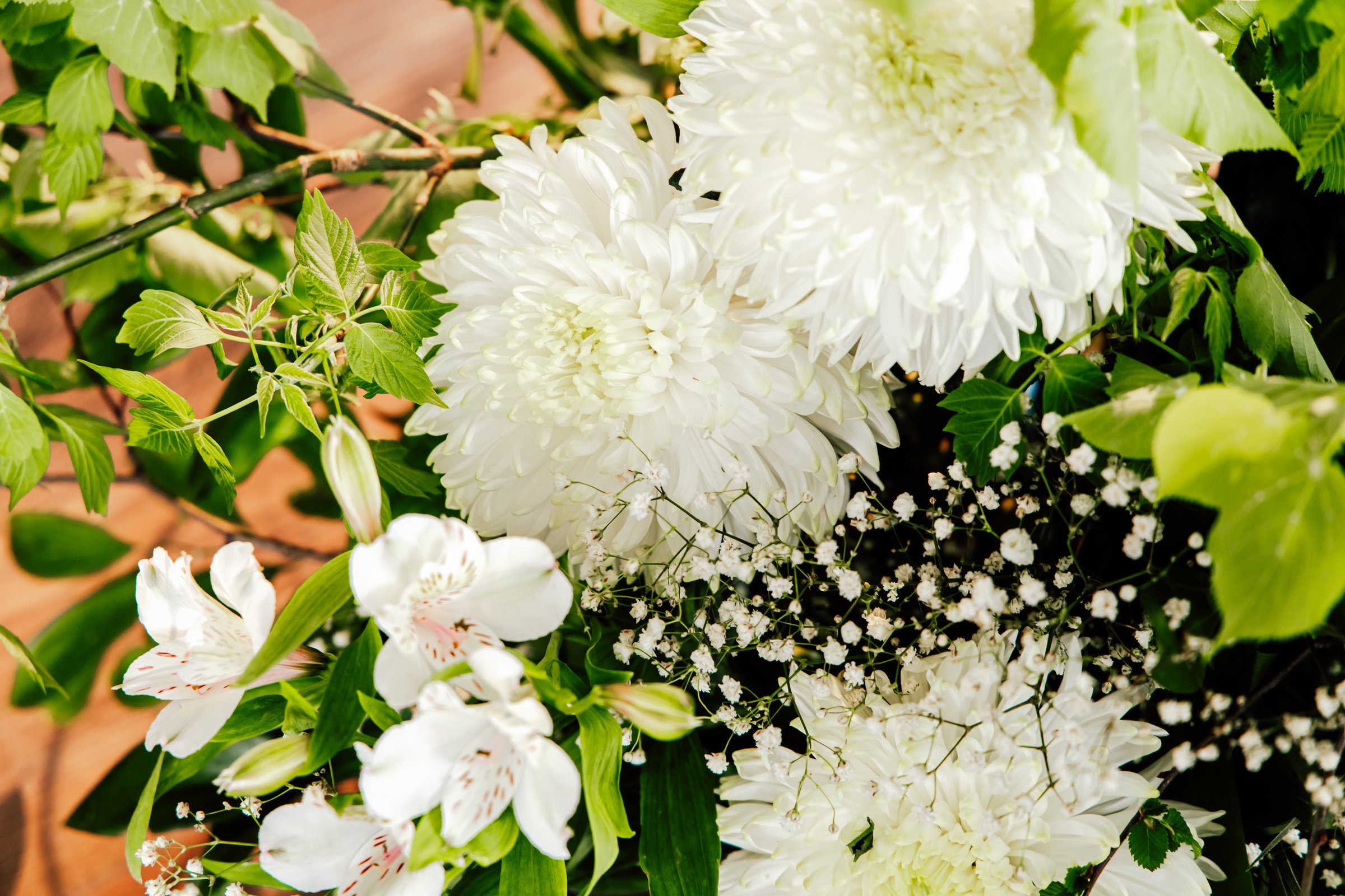 A collection of white flowers featuring chrysanthemums and delicate greenery brightens a wooden surface.