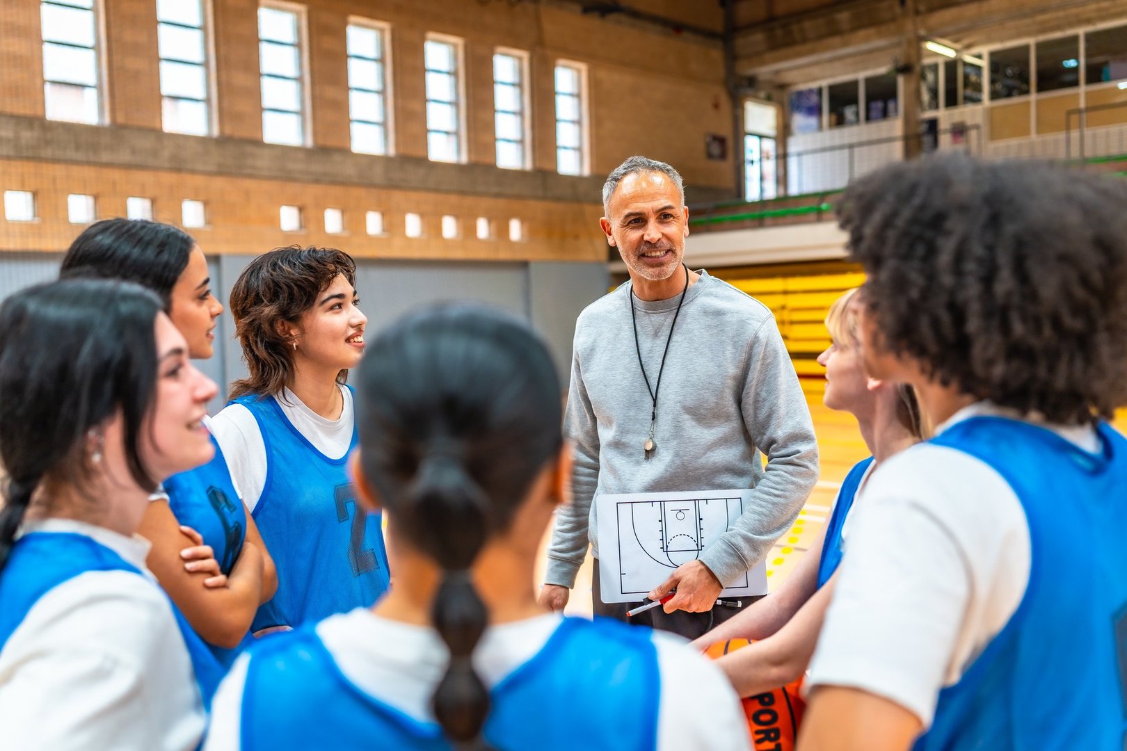 Basketball coach holding clipboard and explaining game strategy to a group of female athletes during training in a gymnasium
