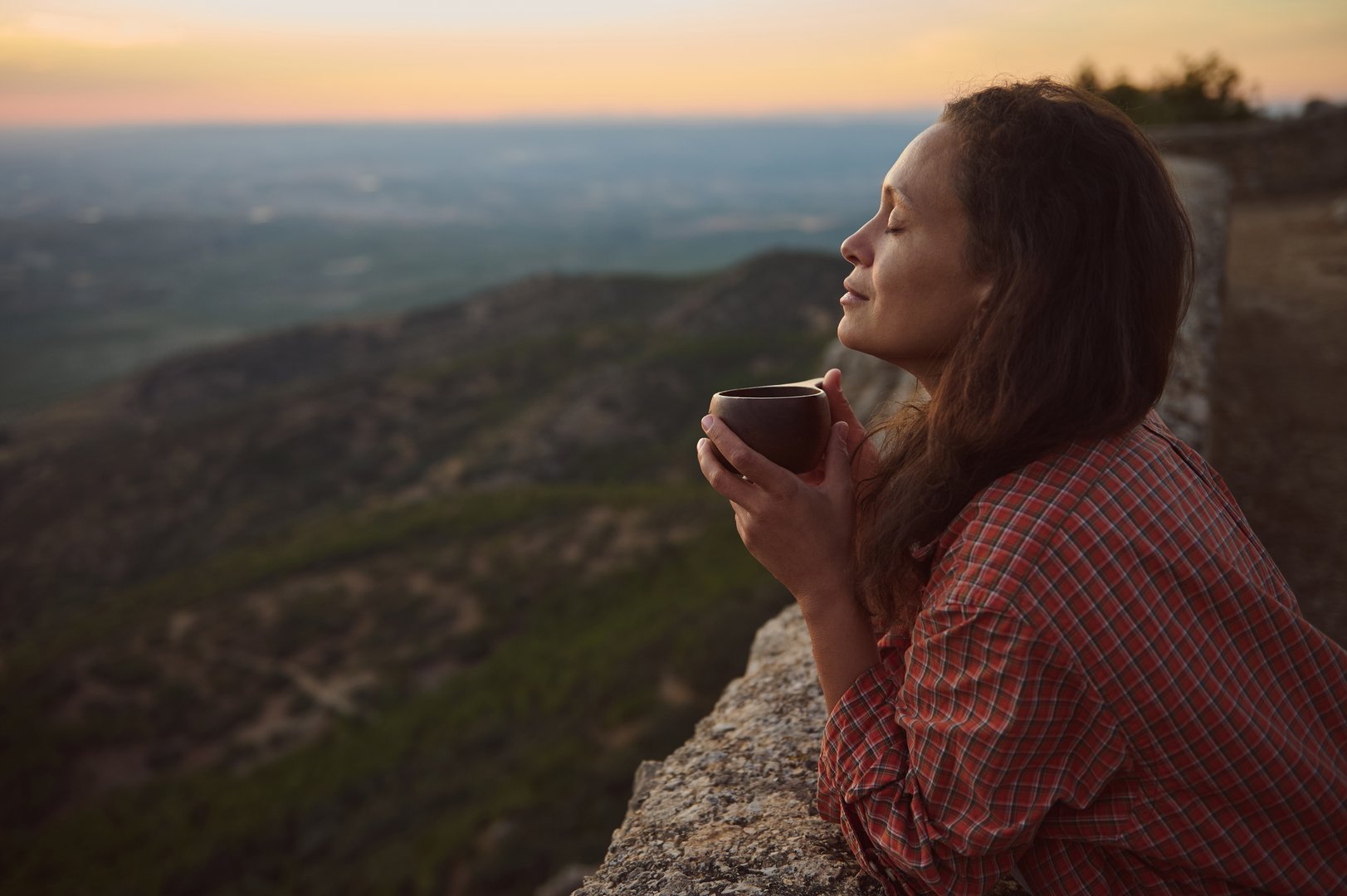 A woman holding a cup of coffee, savoring the peaceful sunrise while overlooking a vast mountainous landscape. Captures calmness and connection with nature.