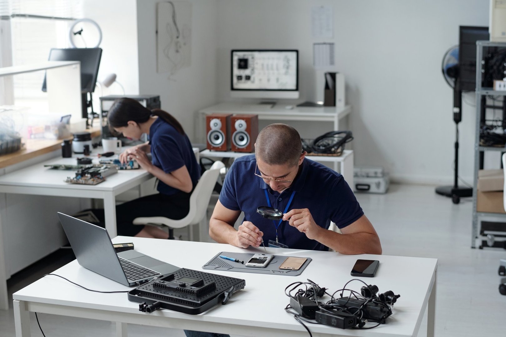 Two technicians focusing on electronics repair in a well-organized workshop. Various tools, computer parts, and devices are neatly arranged on workstations