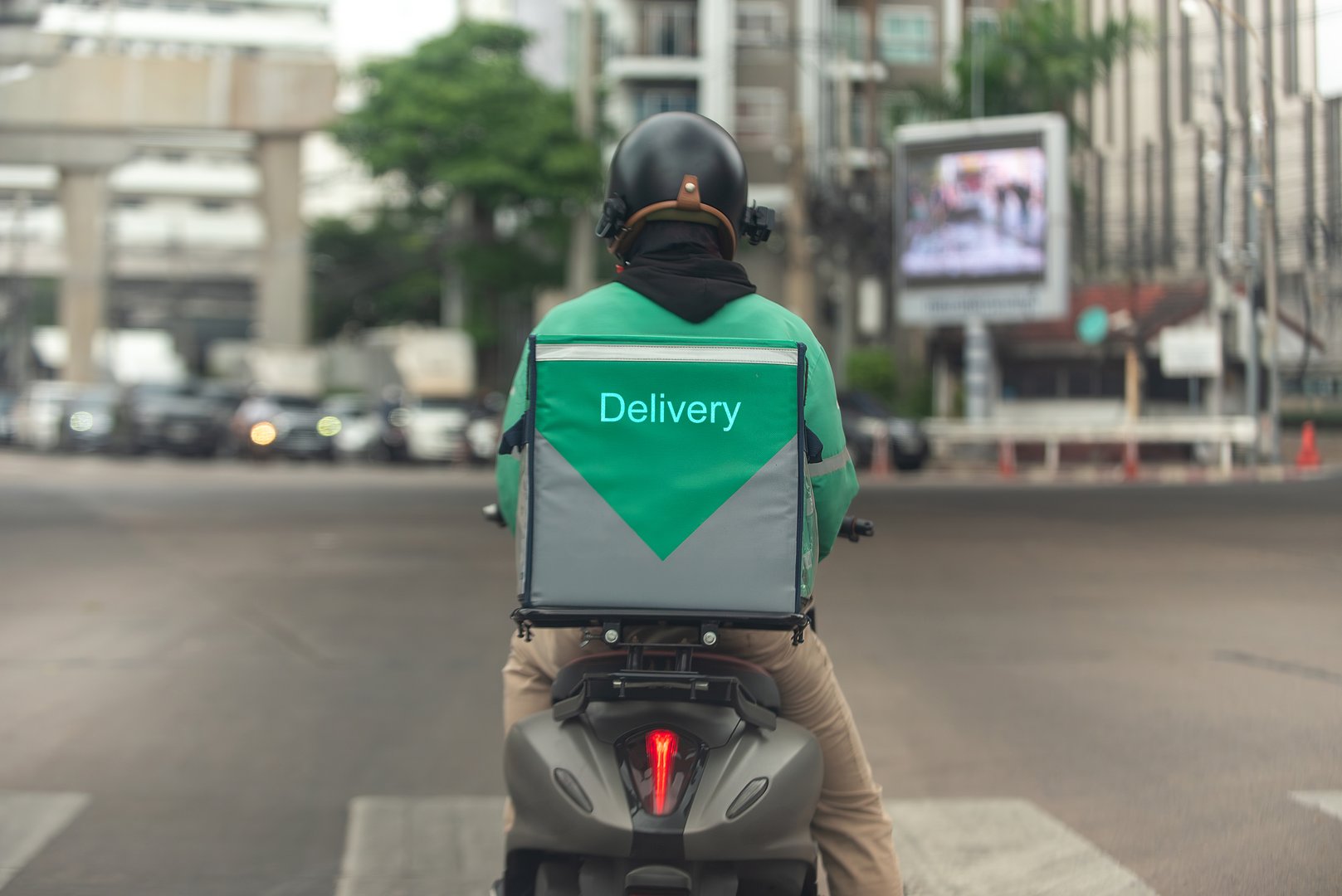Back view of a delivery driver wearing a green uniform and helmet, riding a motorcycle with a large food delivery box on a busy city street. Express logistics concept.