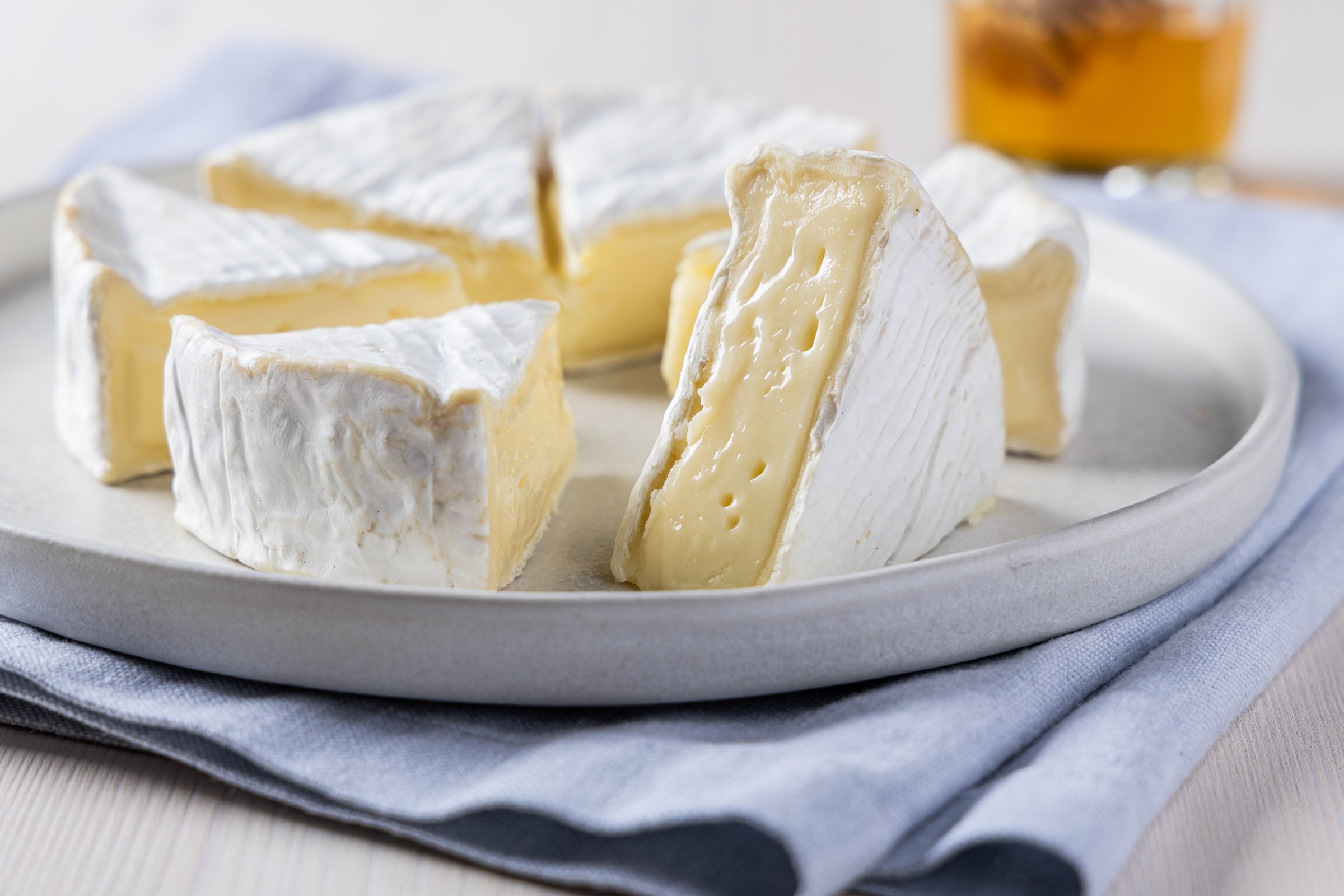 A close-up shot of creamy Camembert cheese on a plate, paired with rustic bread and honey in the background. The image highlights the soft texture and rich flavor of the cheese, perfect for illustrating gourmet food, cheese platters, or culinary blogs.