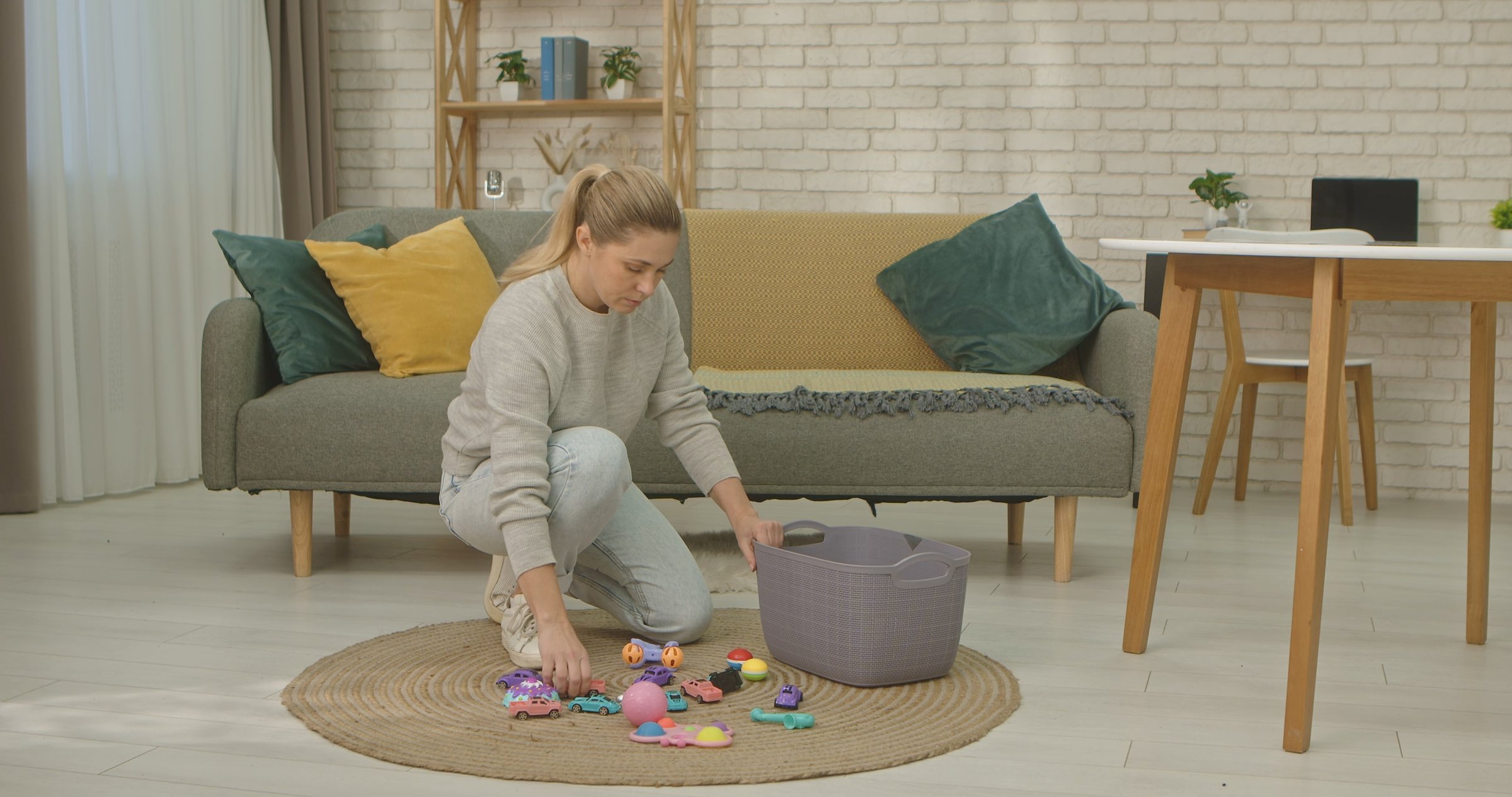 A woman kneels on a rug in a stylish living room, sorting colorful toys into a basket. The room features a modern sofa and decor.