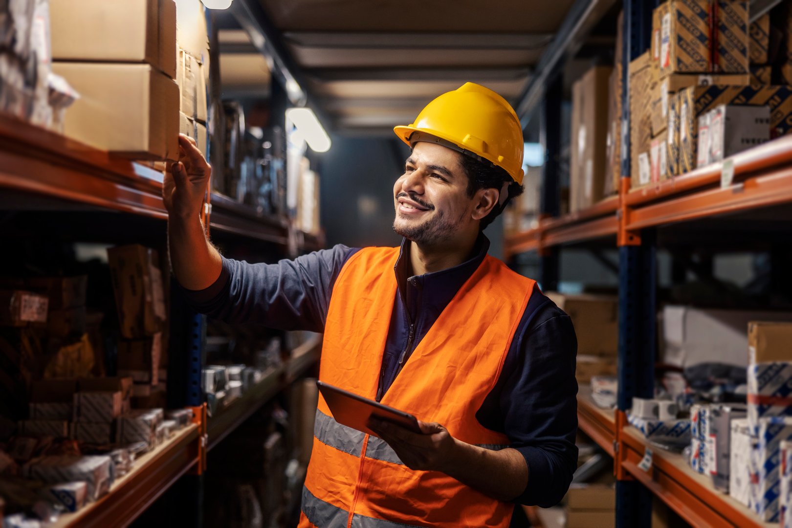 Happy multicultural industry worker with tablet in hands inventorying shipment on shelf at storage.