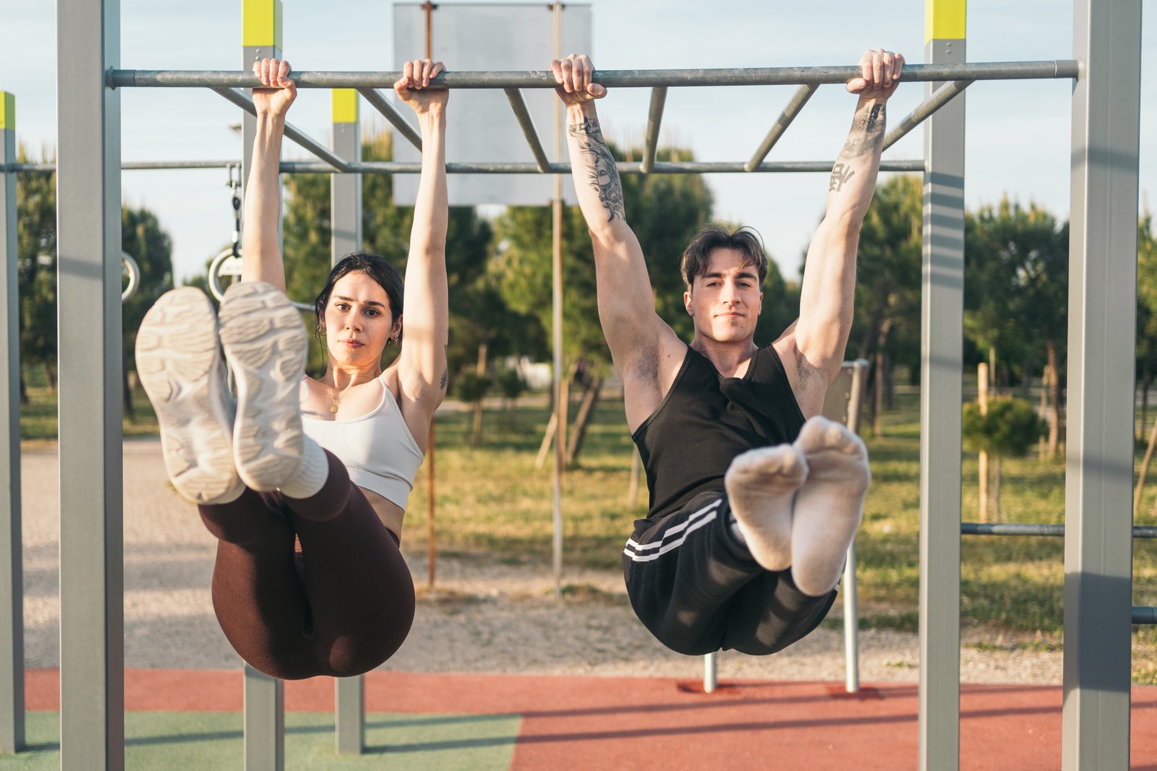 Fit man and woman doing hanged l-sit on outdoor fitness equipment in a park setting