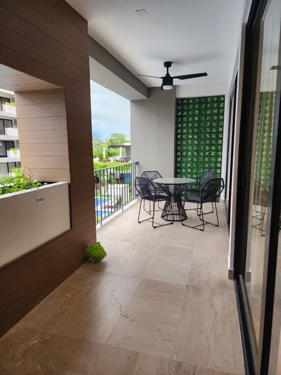 Balcony with a table, four chairs, ceiling fan, and decorative green wall, overlooking a landscaped view.