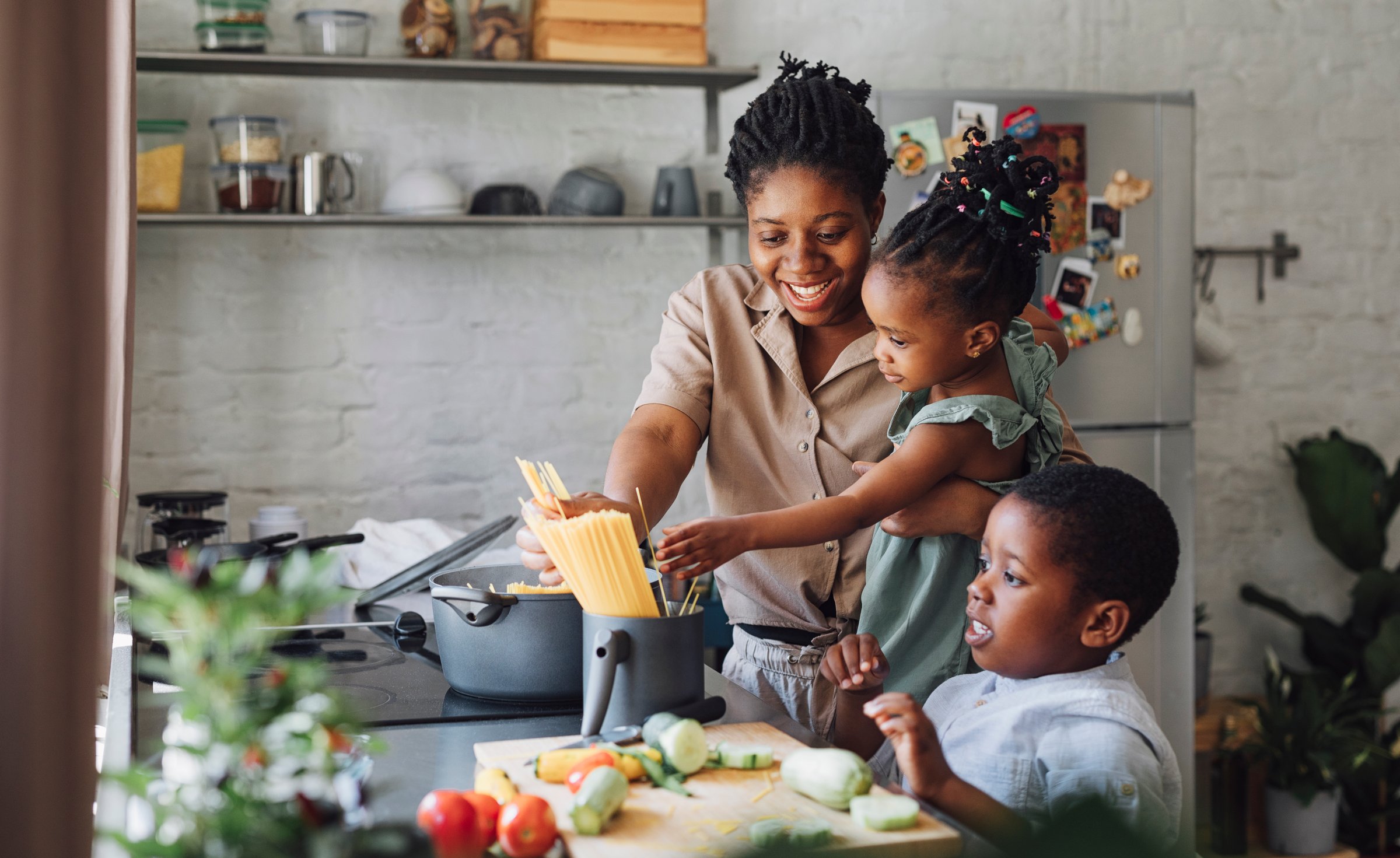 Happy African American mother, daughter and son preparing spaghetti and vegetables for lunch over a kitchen sink.