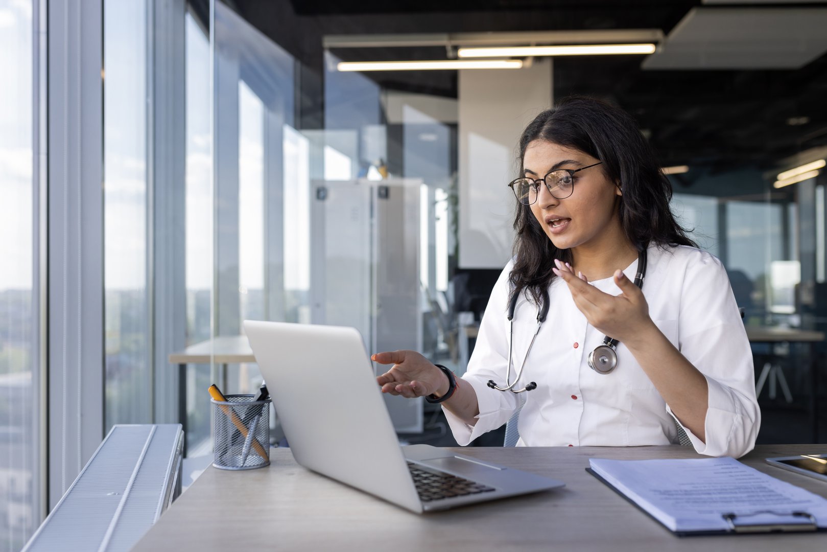 Doctor engaging in virtual consultation using laptop in bright office setting. Professional woman wearing stethoscope, communicating effectively during remote medical appointment.
