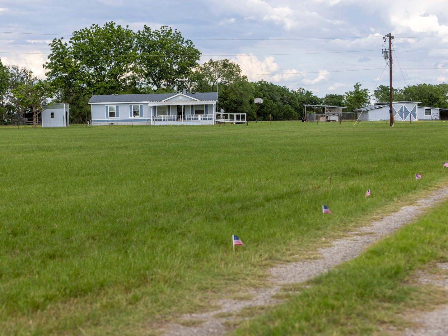 A rural landscape with a white house, a large green field, small American flags lining a path, and some utility lines.