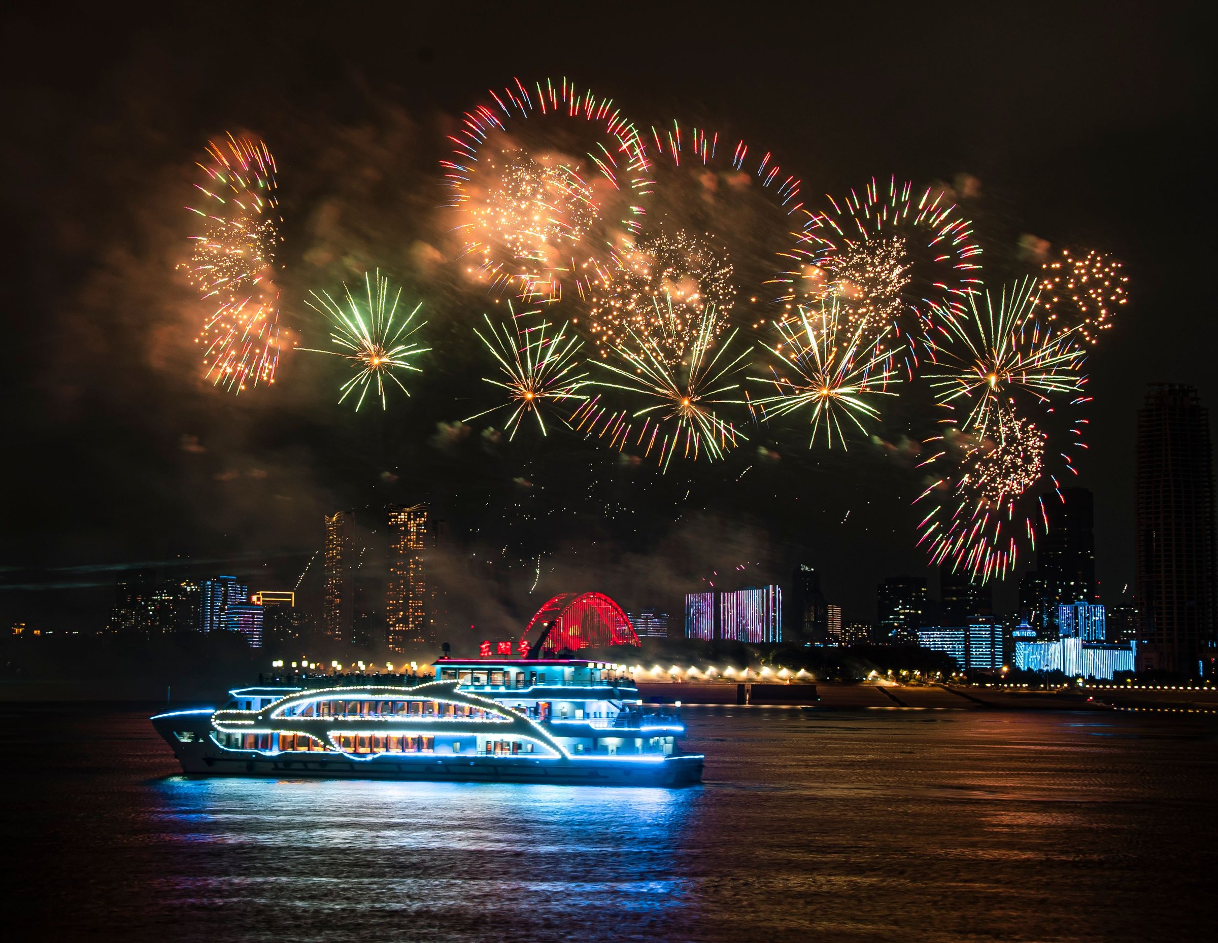 A cruise ship illuminated with blue lights sails on a river with a vibrant fireworks display in the night sky, city skyline in the background.