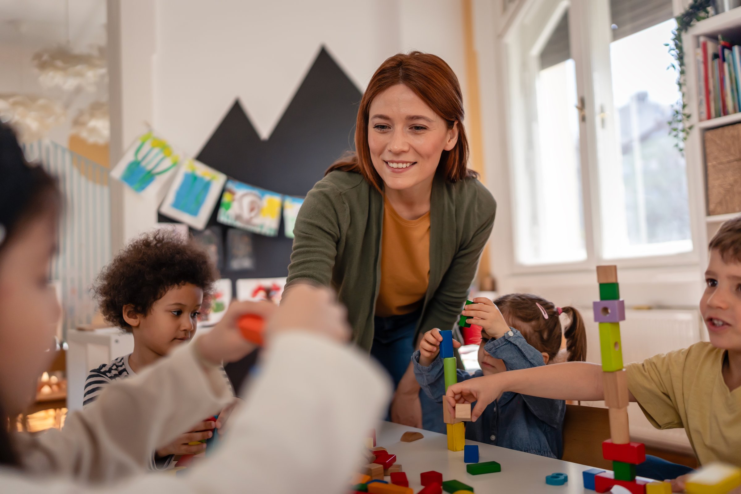 Smiling teacher interacts with diverse group of young children playing with colorful blocks in a bright, cheerful classroom setting.