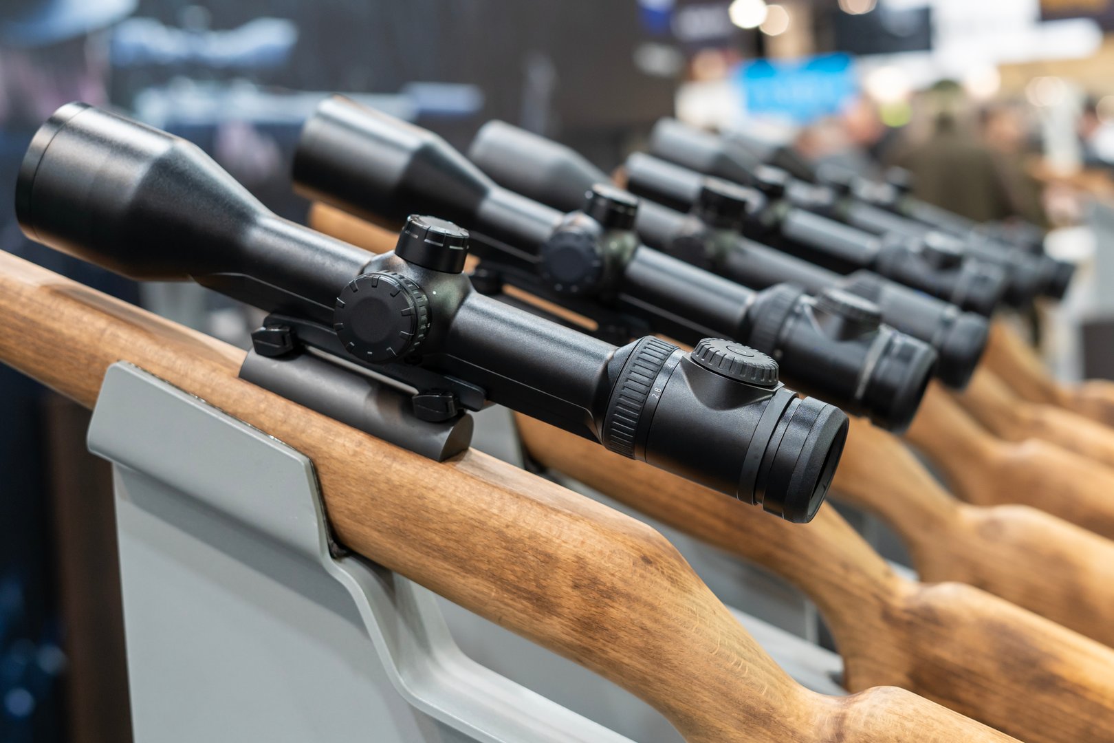 Several wooden rifles equipped with black optical scopes are neatly lined up on a display stand at a trade show or firearms exhibition. The image focuses on the precision scopes, highlighting modern shooting equipment in a professional setting.