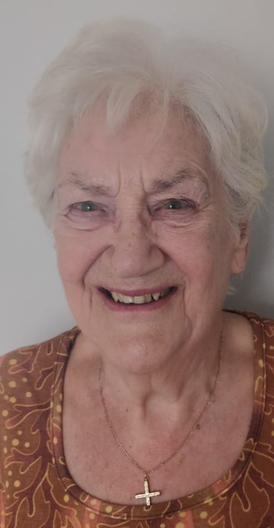 Elderly woman with white hair smiling, wearing a brown patterned top and a necklace with a cross pendant.