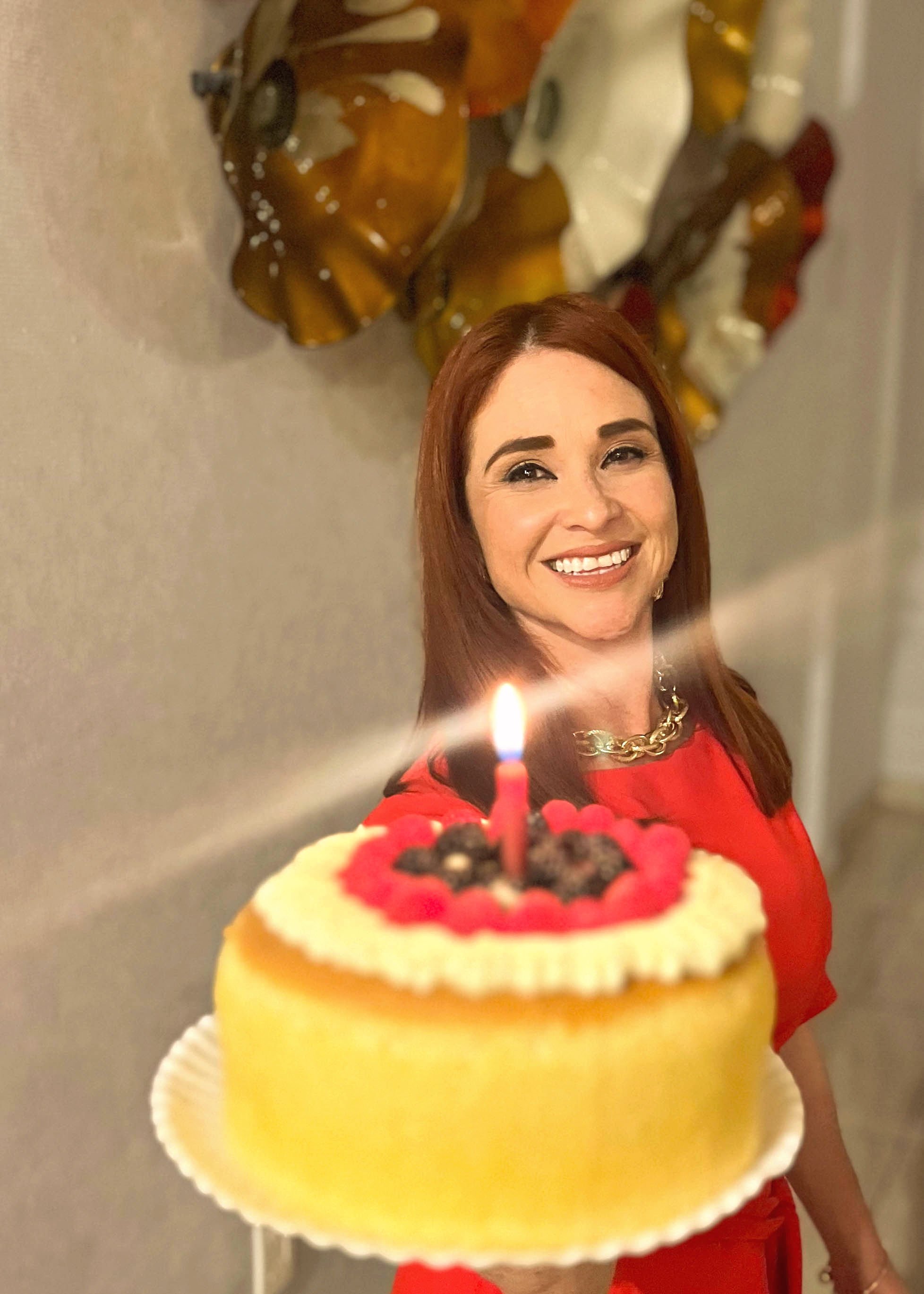 Woman smiling and holding a lit birthday cake with colorful decorations in a room.