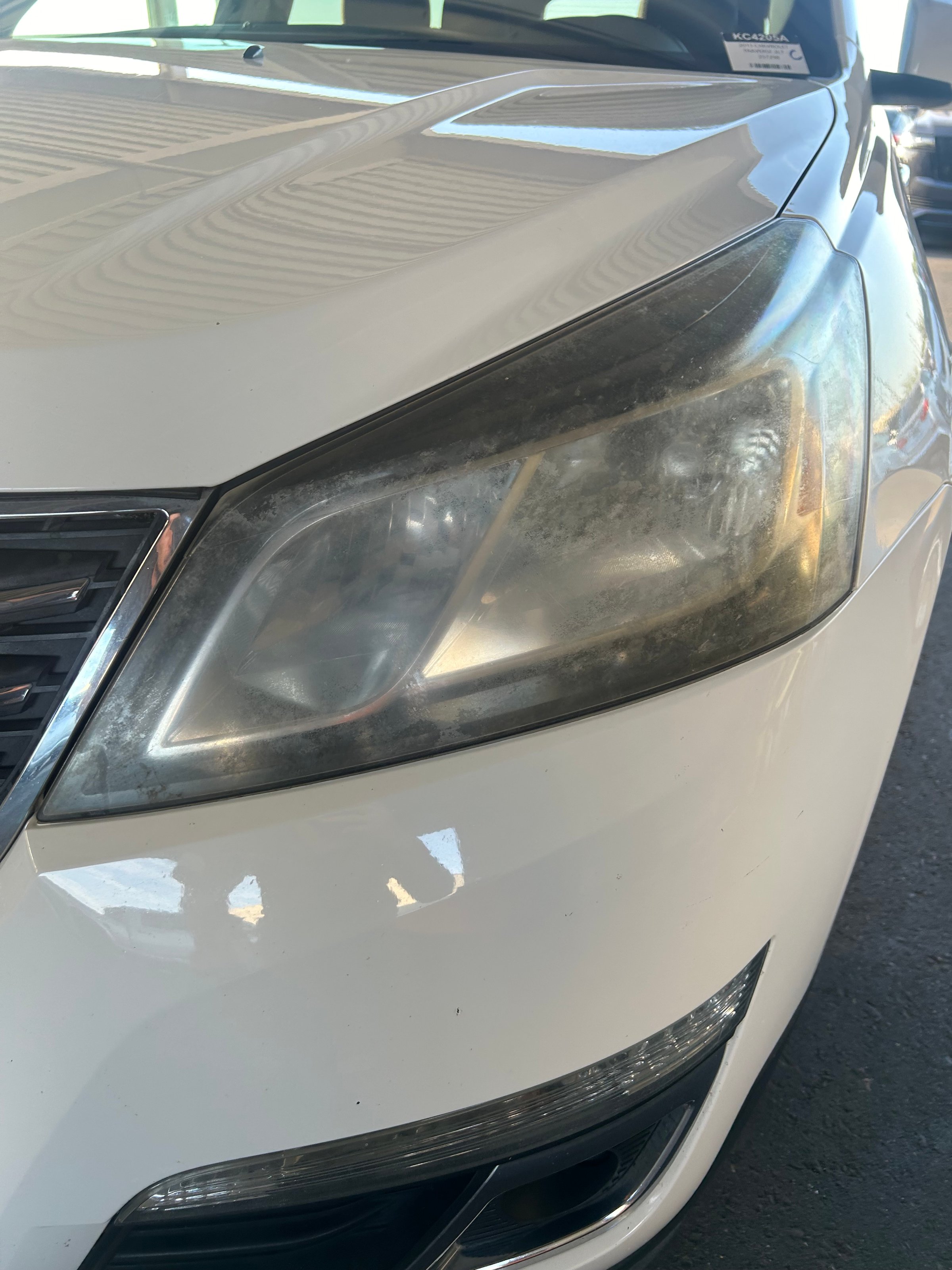 Close-up of a white car's front left headlight, showing signs of age and wear, parked in a covered area.