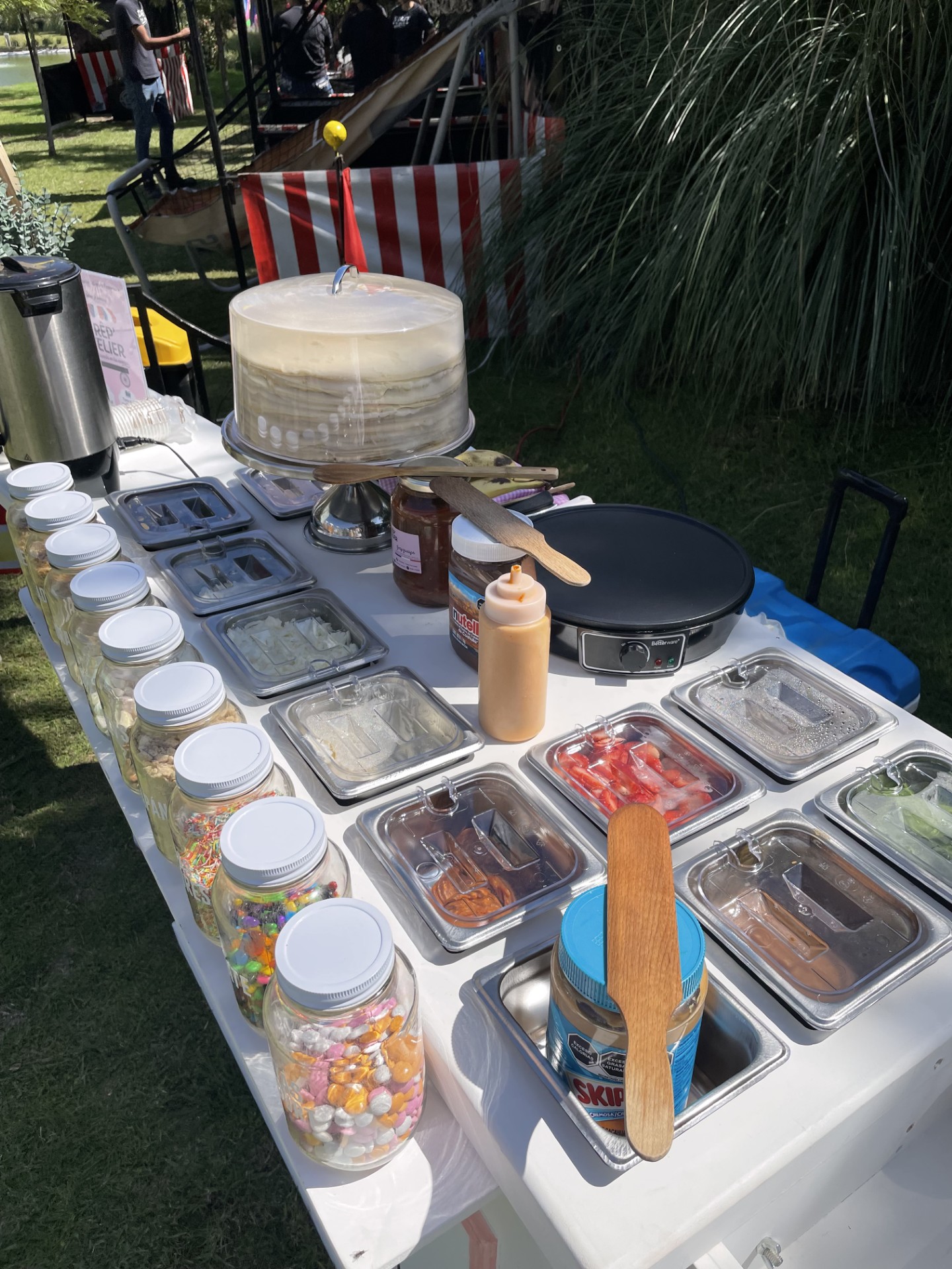 A table with a crepe-making station, various toppings in jars and trays, and a cake displayed under a dome on a sunny day.