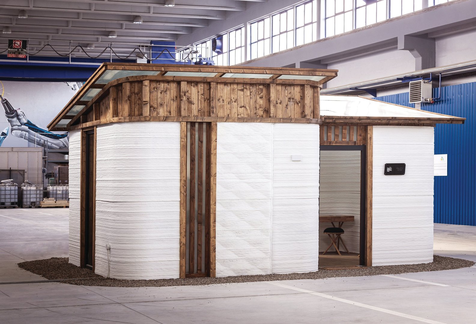 A small, modern 3D-printed house with wood accents, inside an industrial building. It has a flat roof and white textured walls.