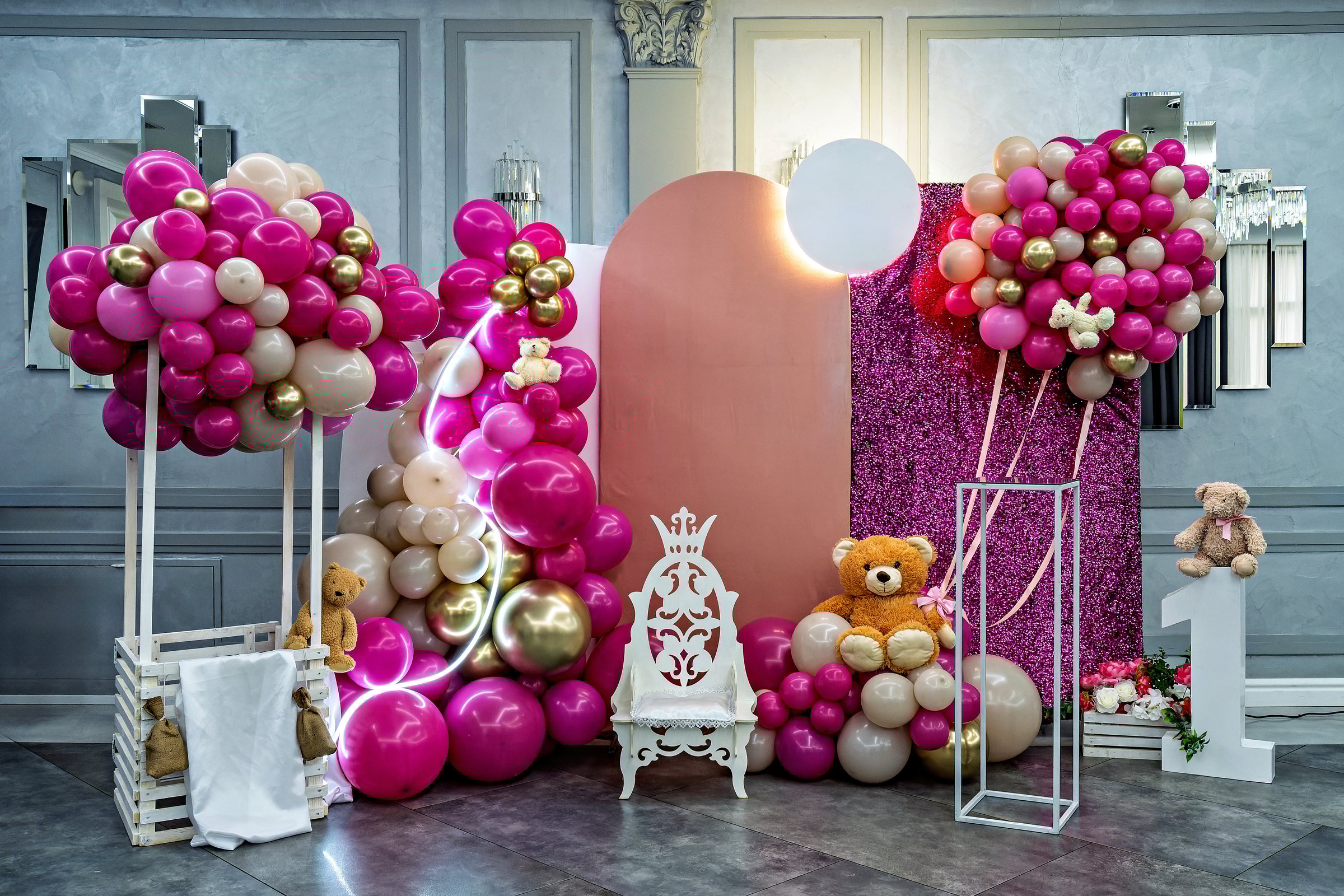 A room decorated with a large pink balloon arch a pink chair and a teddy bear with a sign that says Nika and a number 1 on the right