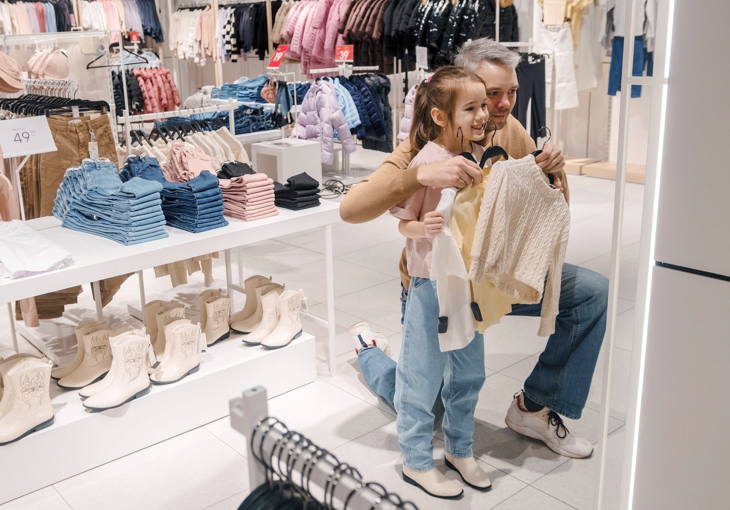 A father helps his excited daughter choose clothes in a brightly lit mall, surrounded by folded clothing and footwear.