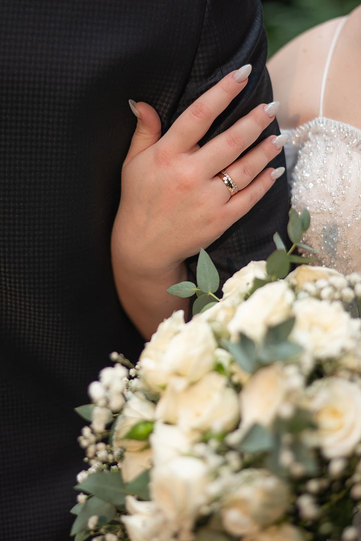 bride hugs groom and holds wedding bouquet in hands behind back close-up golden engagement rings are visible. couple at wedding ceremony. white flowers and dress