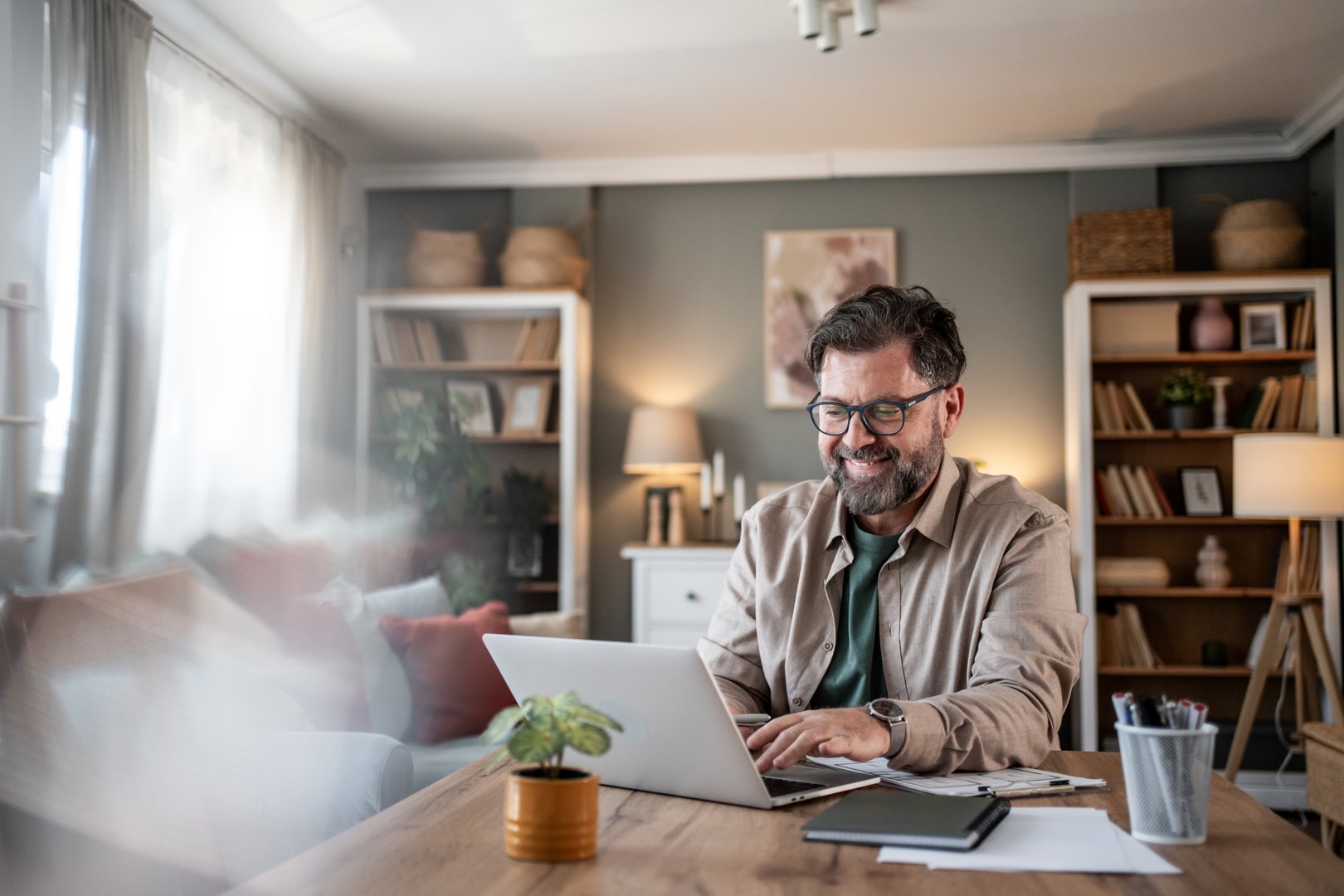 Happy mid-adult man wearing eyeglasses, sitting at a wooden table in a cozy home office, working remotely on his laptop and smiling while enjoying the comfort of his space