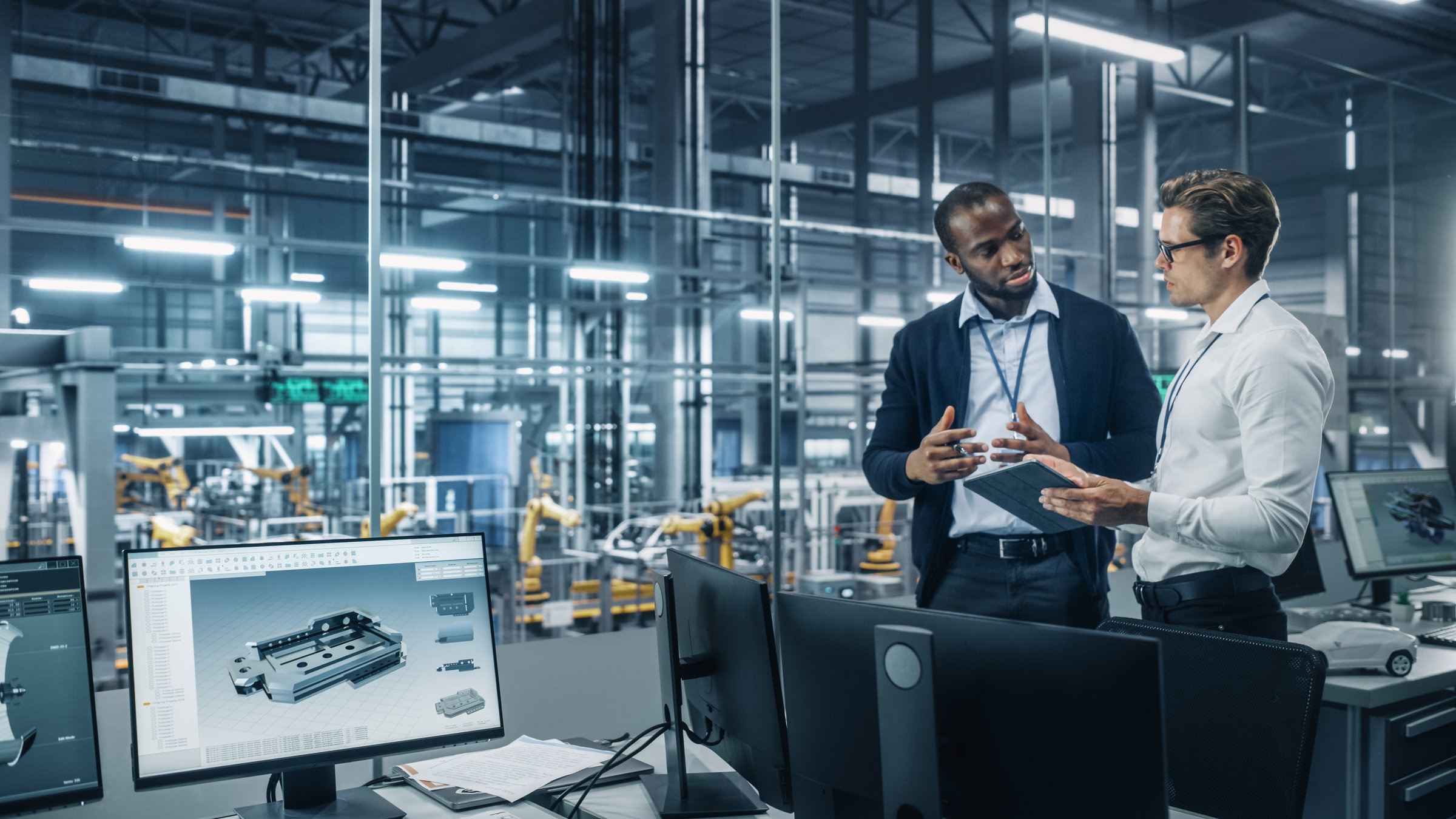 Two Diverse Automotive Industrial Engineers Talking About Vehicle Production while Standing in Office at a Car Assembly Plant. Industrial Specialists Discuss Work Projects on Tablet Computer.