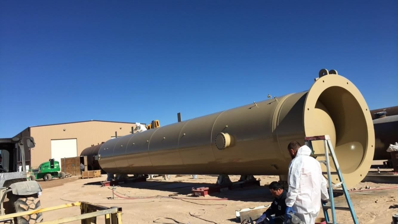 Large beige industrial cylinder on blocks outdoors with workers in protective suits nearby, building in background.