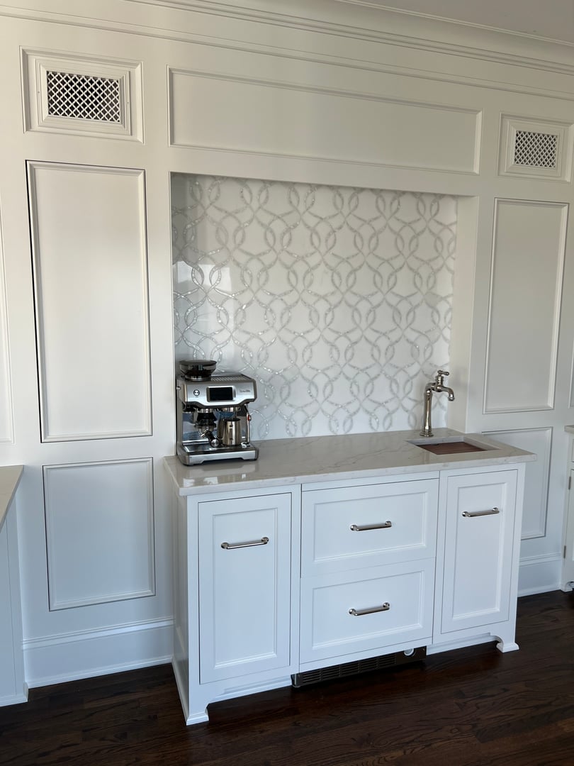 White kitchen setup with a coffee maker, sink, and patterned backsplash on a marble countertop, set against paneled walls.