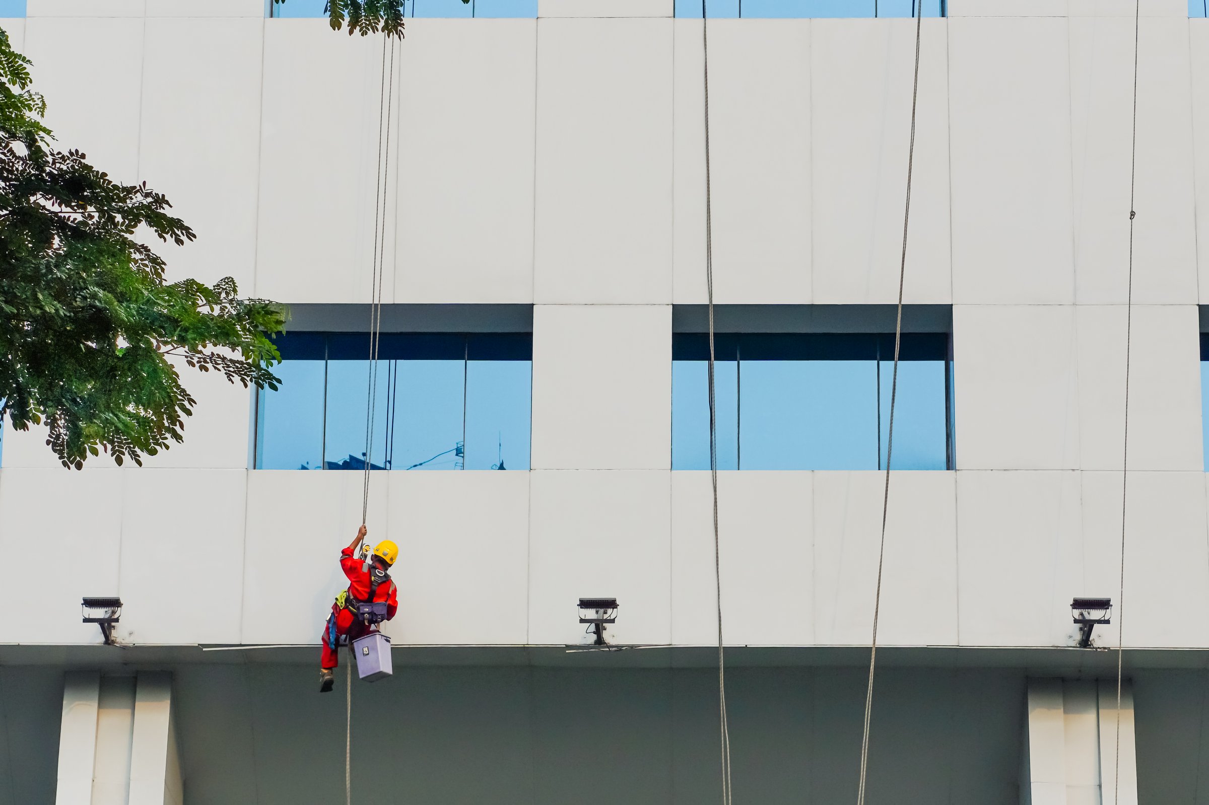 Serpong, Indonesia - July 21, 2024: A professional worker in full protective gear is cleaning the glass of a high-rise building using a hanging rope system. Seen hanging in front of a modern building facade with blue windows.