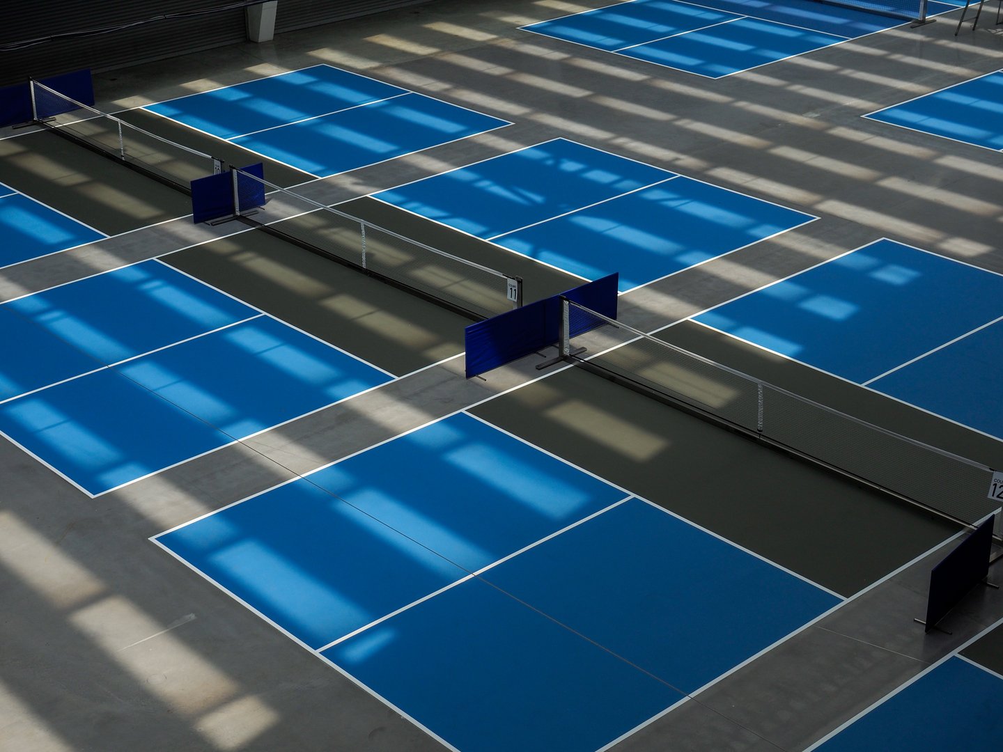 Pickleball court indoor with blue service area and dark grey area known as the kitchen or non volley area with some light and shadows in play.