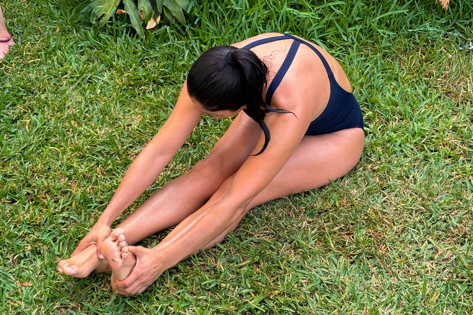 A person in a black swimsuit stretching forward to touch their toes while sitting on grass.