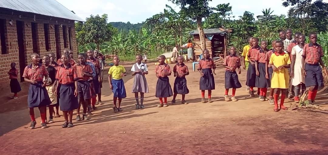 Children in school uniforms standing outside on dirt ground, with greenery and a building in the background.