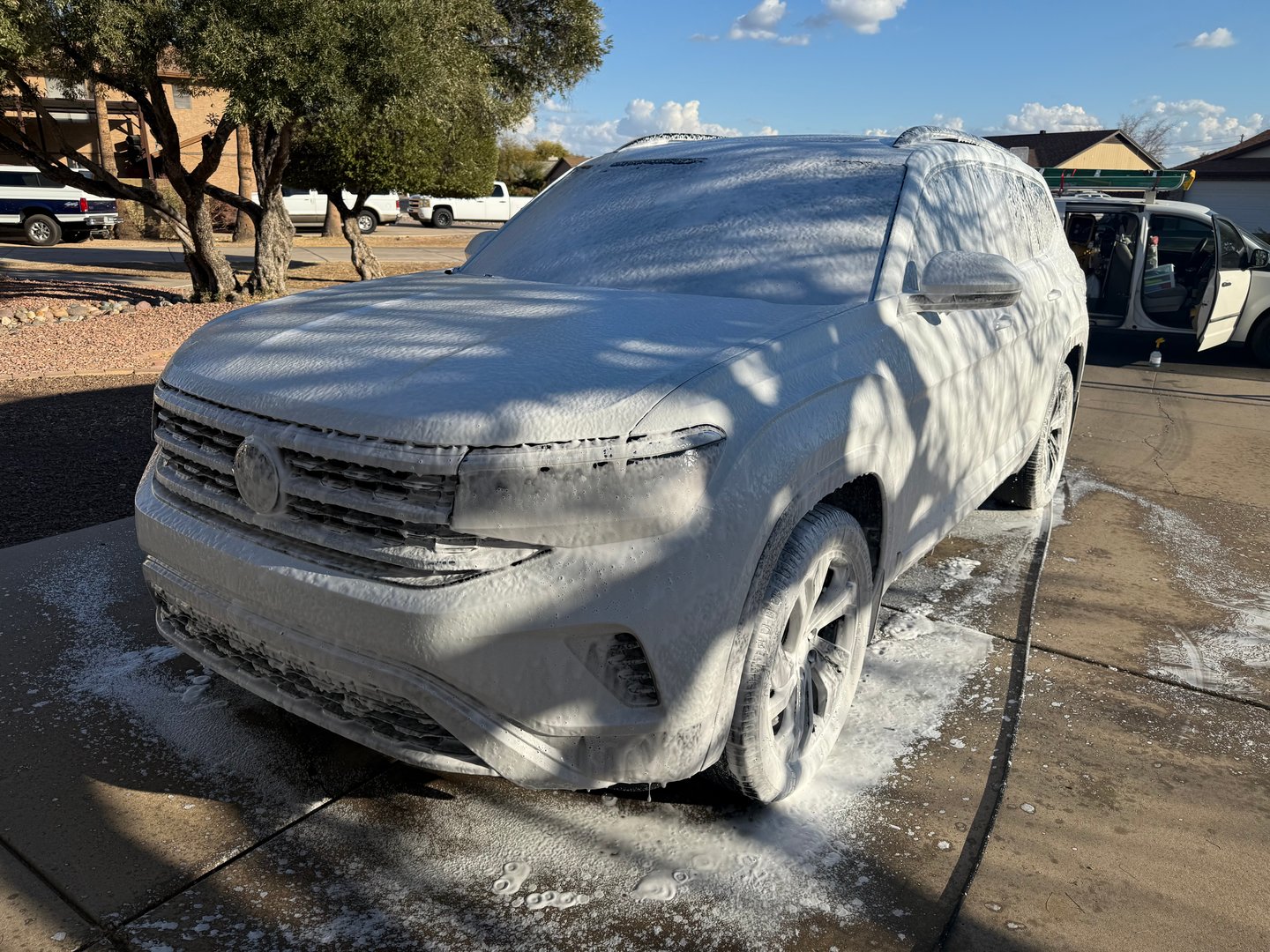 SUV covered in white foam during a car wash, parked in a driveway under tree shade on a sunny day.