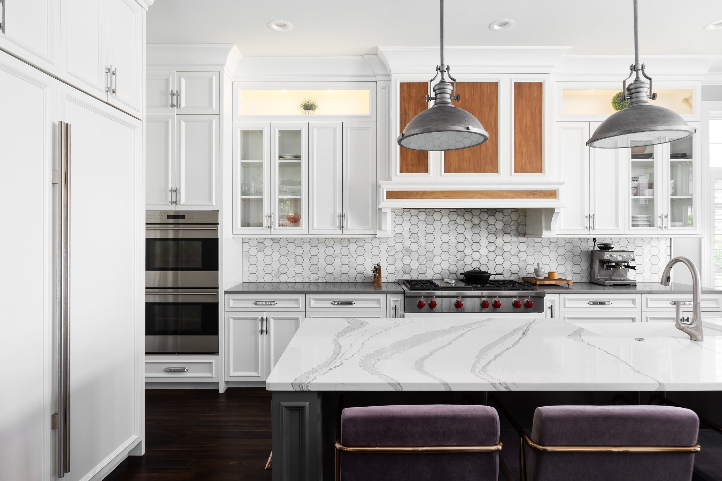 A kitchen detail with metal lights hanging above a large marble island, stainless faucet on a white farmhouse sink, and a marble hexagon tiled backsplash with white cabinets. No brands or labels.