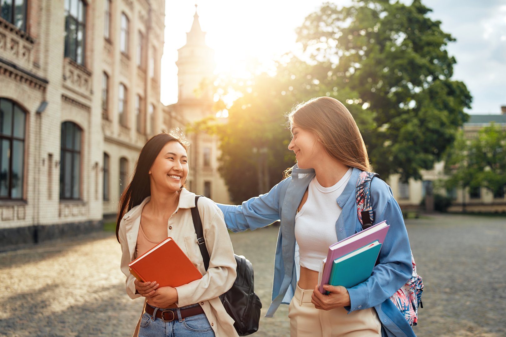 On a beautiful sunny day, two university students stroll through campus, chatting and carrying books. They seem joyful and engaged, enjoying the outdoors amid the academic setting