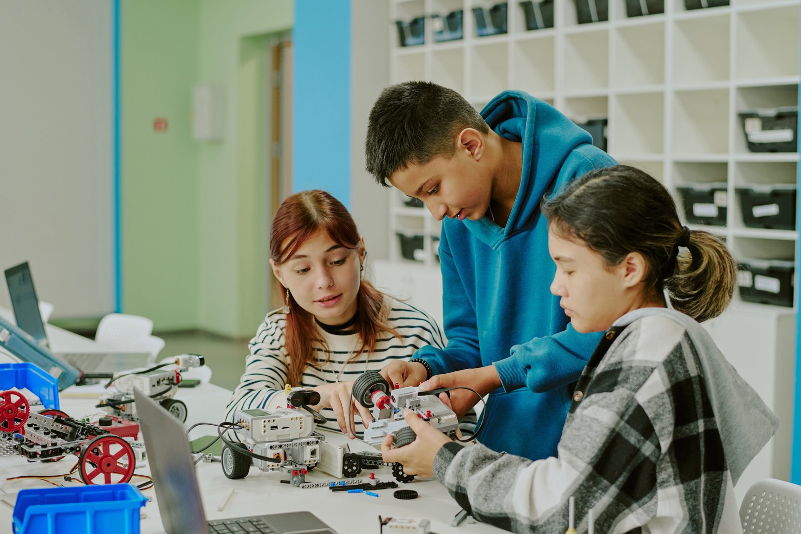 School boy in blue hoodie helping with robot wheels while girls watching process at table