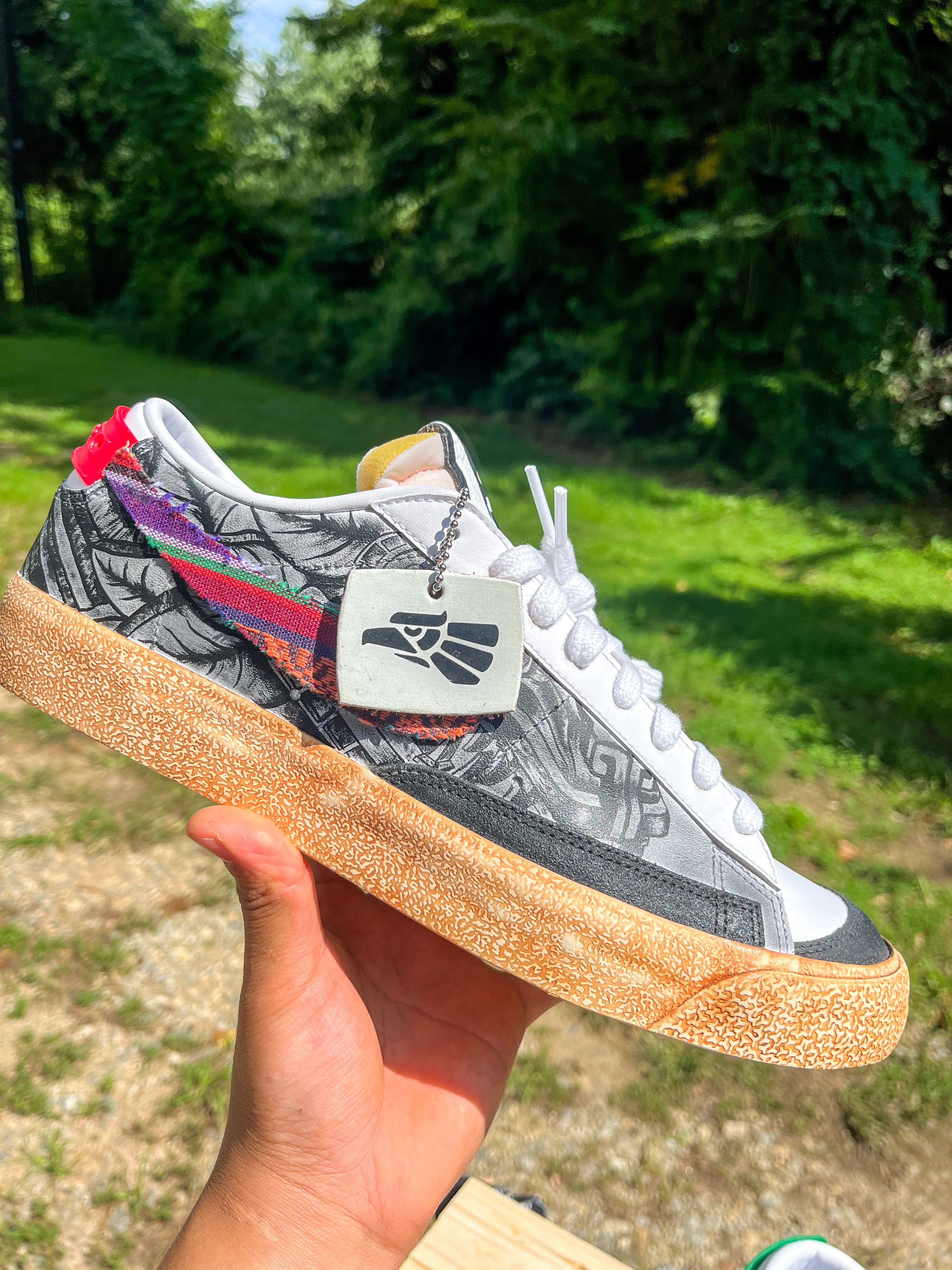 Hand holding a sneaker with colorful accents and a textured sole, outdoors with greenery in the background.