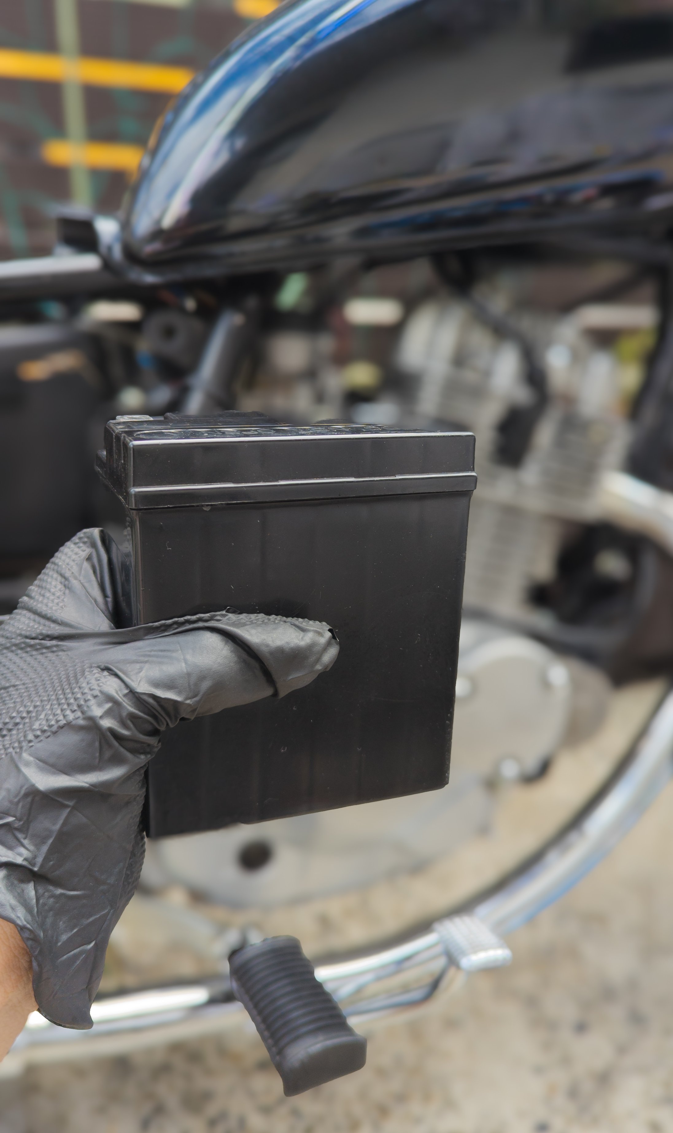 A left hand wearing a black disposable latex glove holding a motorcycle black battery.