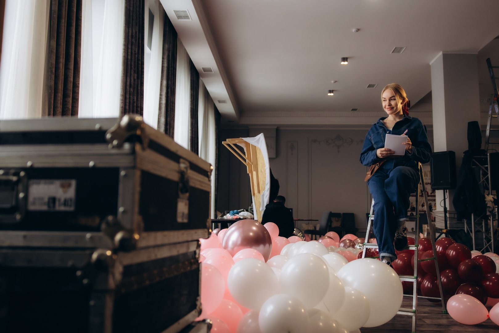 Event planner woman sitting on a ladder, writing a checklist for decorating a spacious room with many balloons
