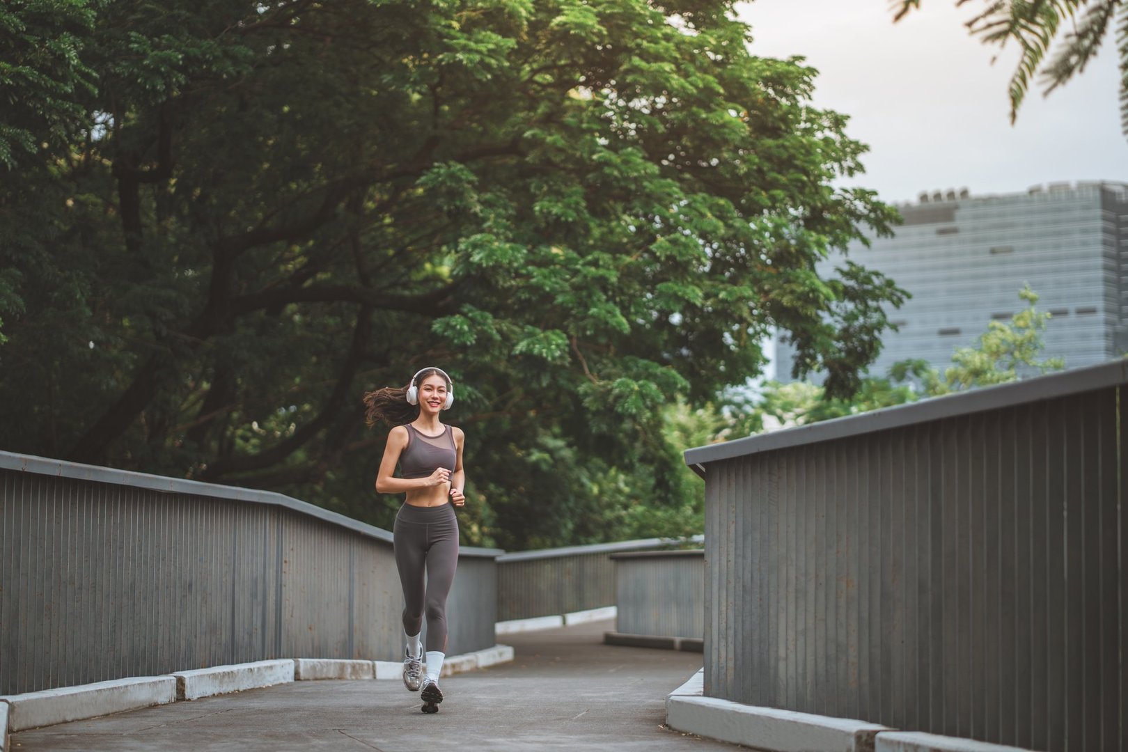 Happy asian female athlete wearing headphones running through shaded city park, active lifestyle, outdoor fitness and wellness concept with space for copy, sportswear and healthy living inspiration