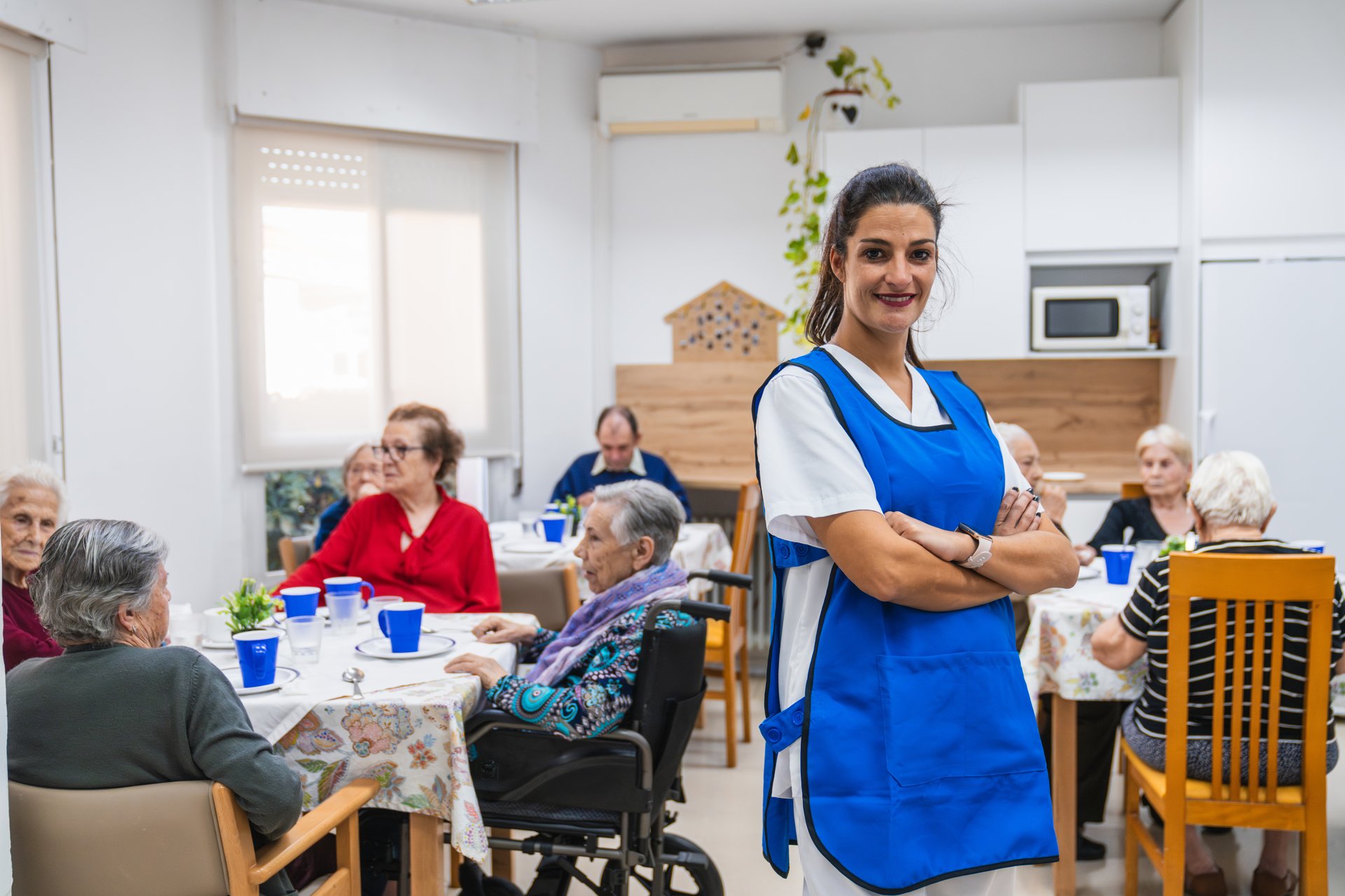 Nurse in a blue apron assisting senior adults enjoying a communal meal in a nursing home dining area, fostering community and care