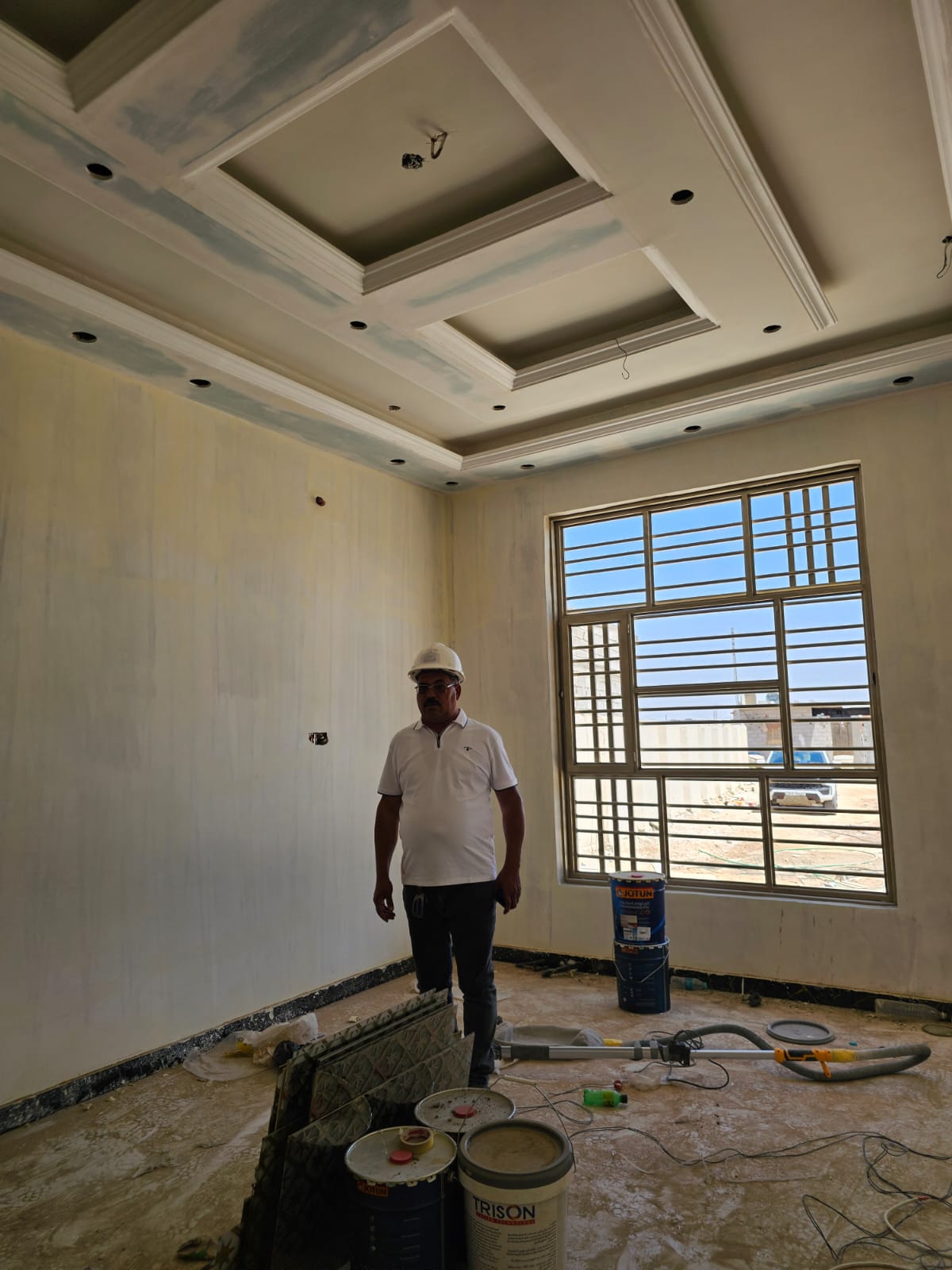 Construction worker in a partially painted room with ceiling lights, standing near paint cans and a large window.
