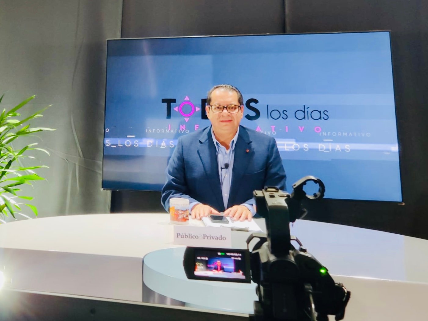 Man in suit hosting a news show at a desk with a camera recording, large screen behind displaying TOS los días.